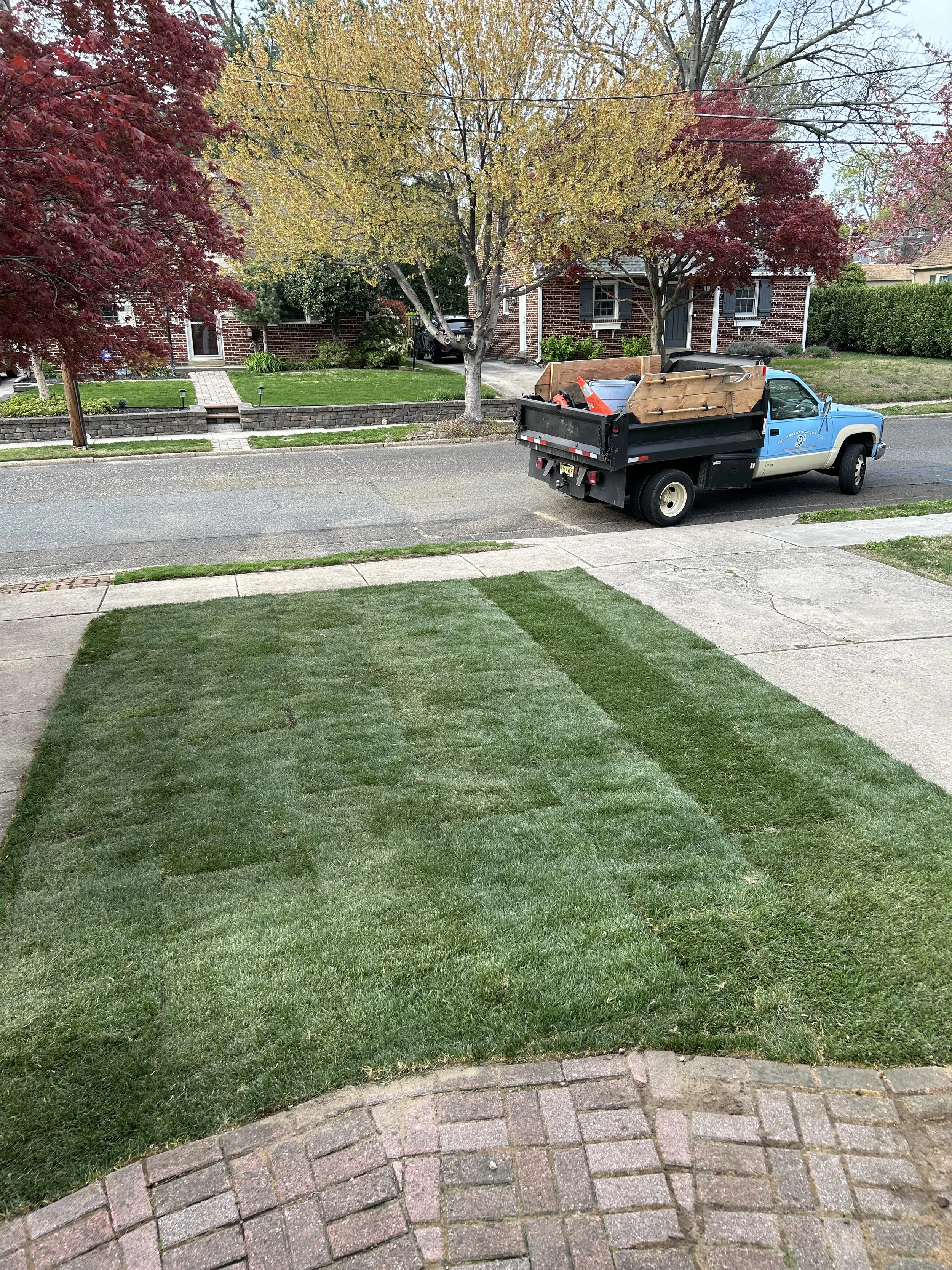 Front yard with green grass, a brick walkway, a tree with yellow and red leaves, and a moving truck parked on the street.