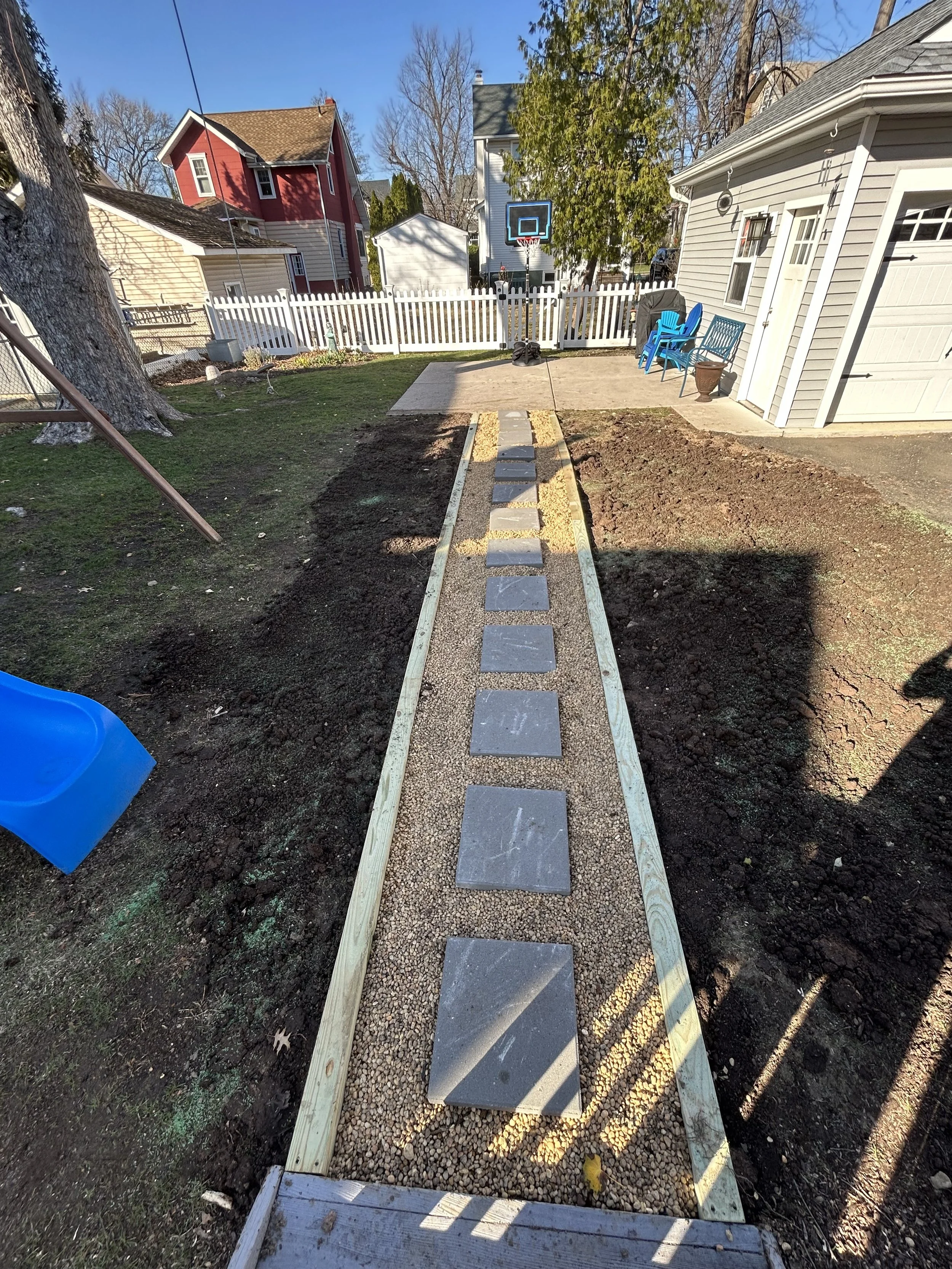 Backyard with a newly constructed stone and concrete pathway leading to a basketball hoop, surrounded by dirt and grass, with outdoor furniture and a white picket fence.