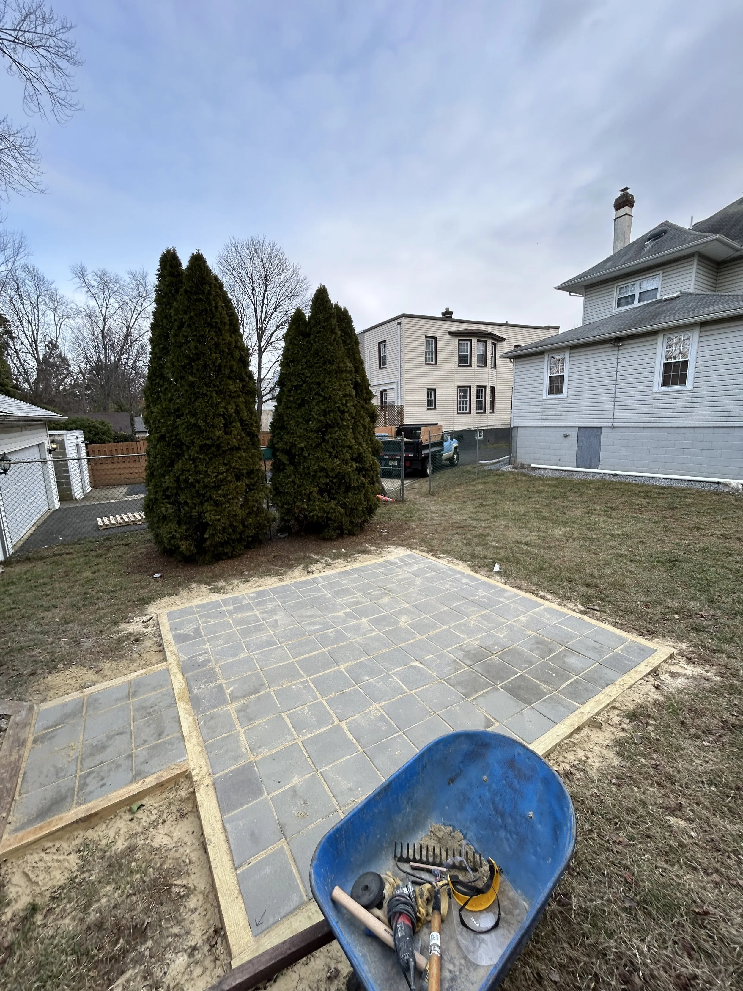 A backyard with a partly paved section, three tall evergreen trees, a wheelbarrow with tools, and a residential house and building in the background.