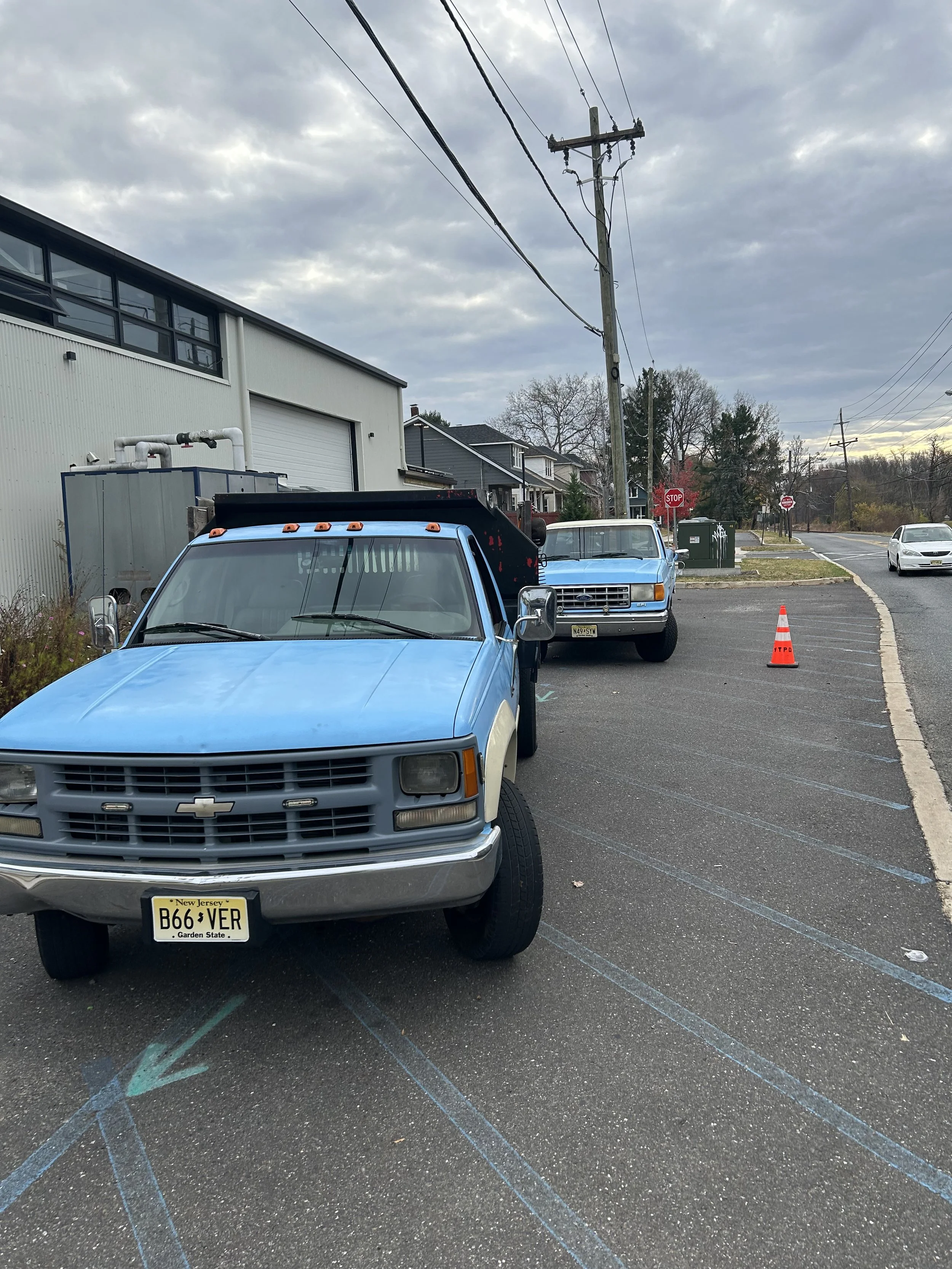 A parking lot with two blue pickup trucks parked, one in the foreground and one further back, with orange traffic cones on the side. There are marked parking spaces, a building with a roller door, some houses, utility poles, and overhead wires under a cloudy sky.