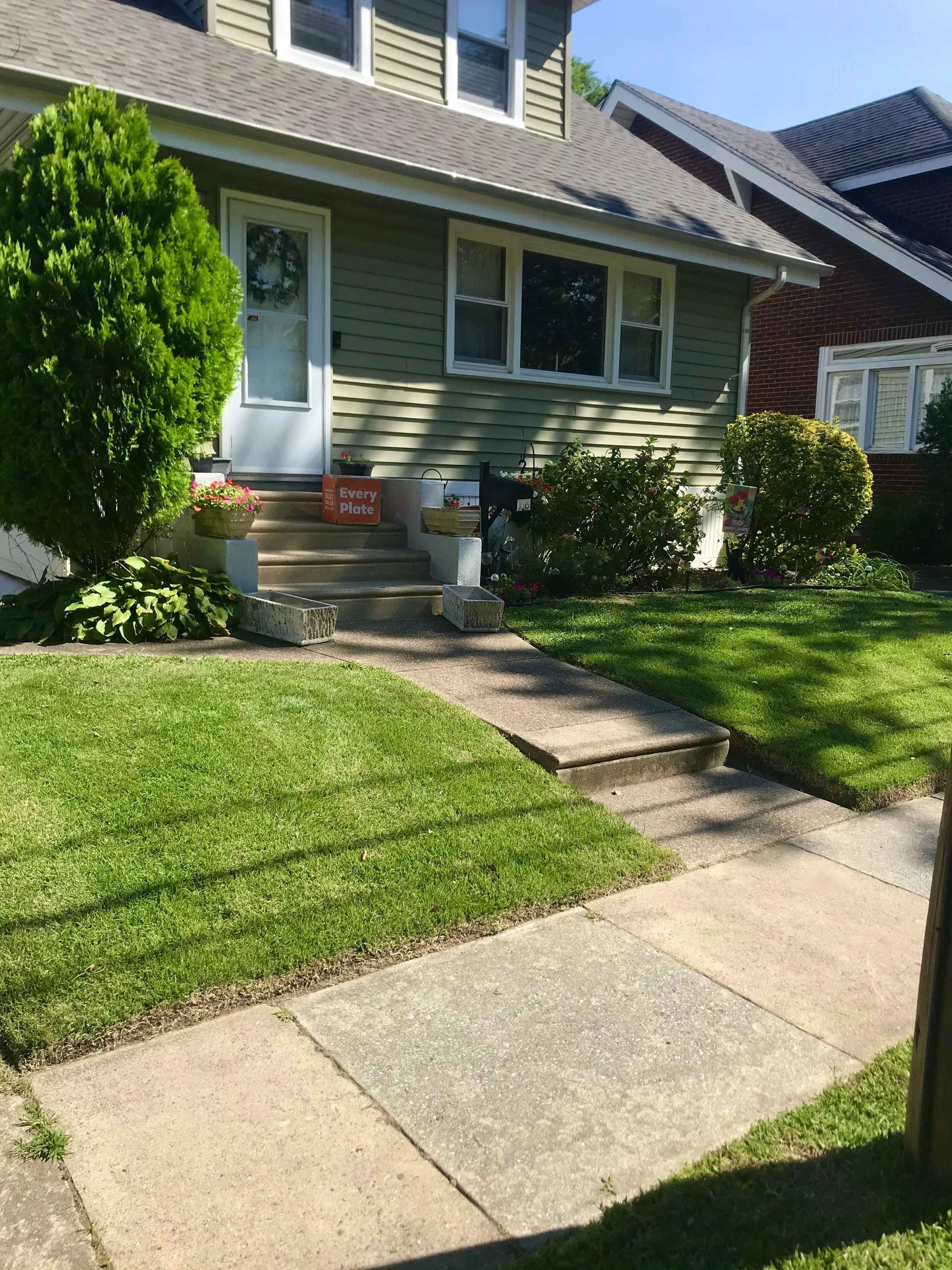 Front yard of a house with green lawn, concrete walkway, bushes, and potted plants near the front door, which has steps and a mailbox.