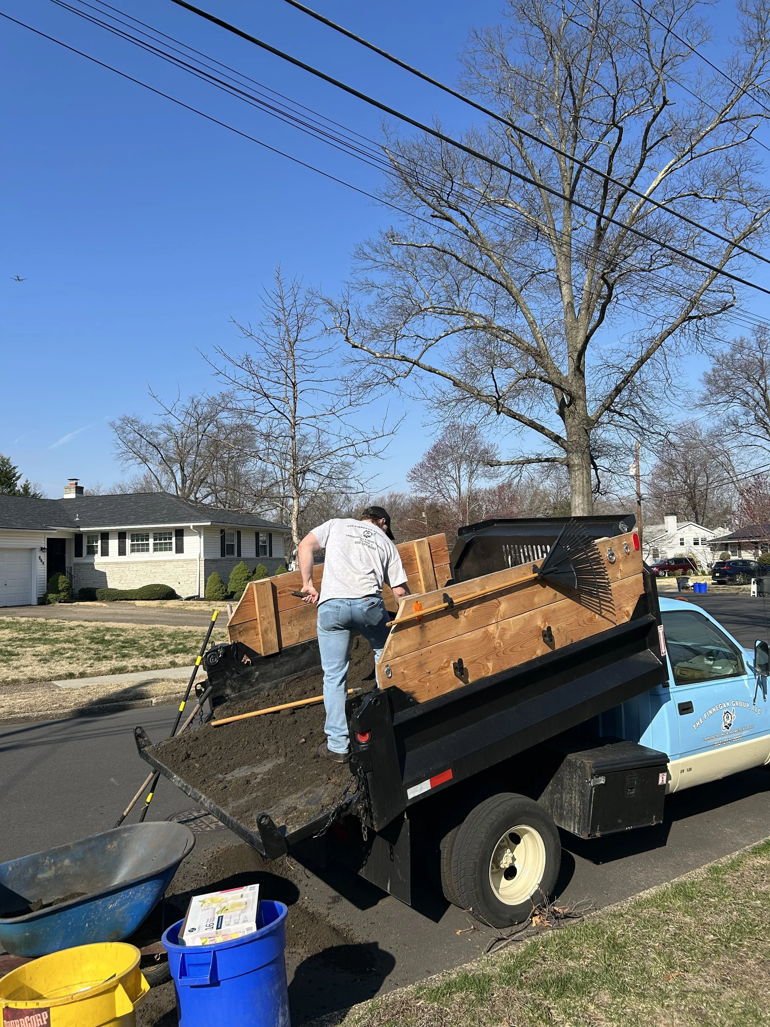 A person is using a small dump truck to unload soil or dirt on a street in a residential neighborhood with trees and houses, under a clear blue sky.
