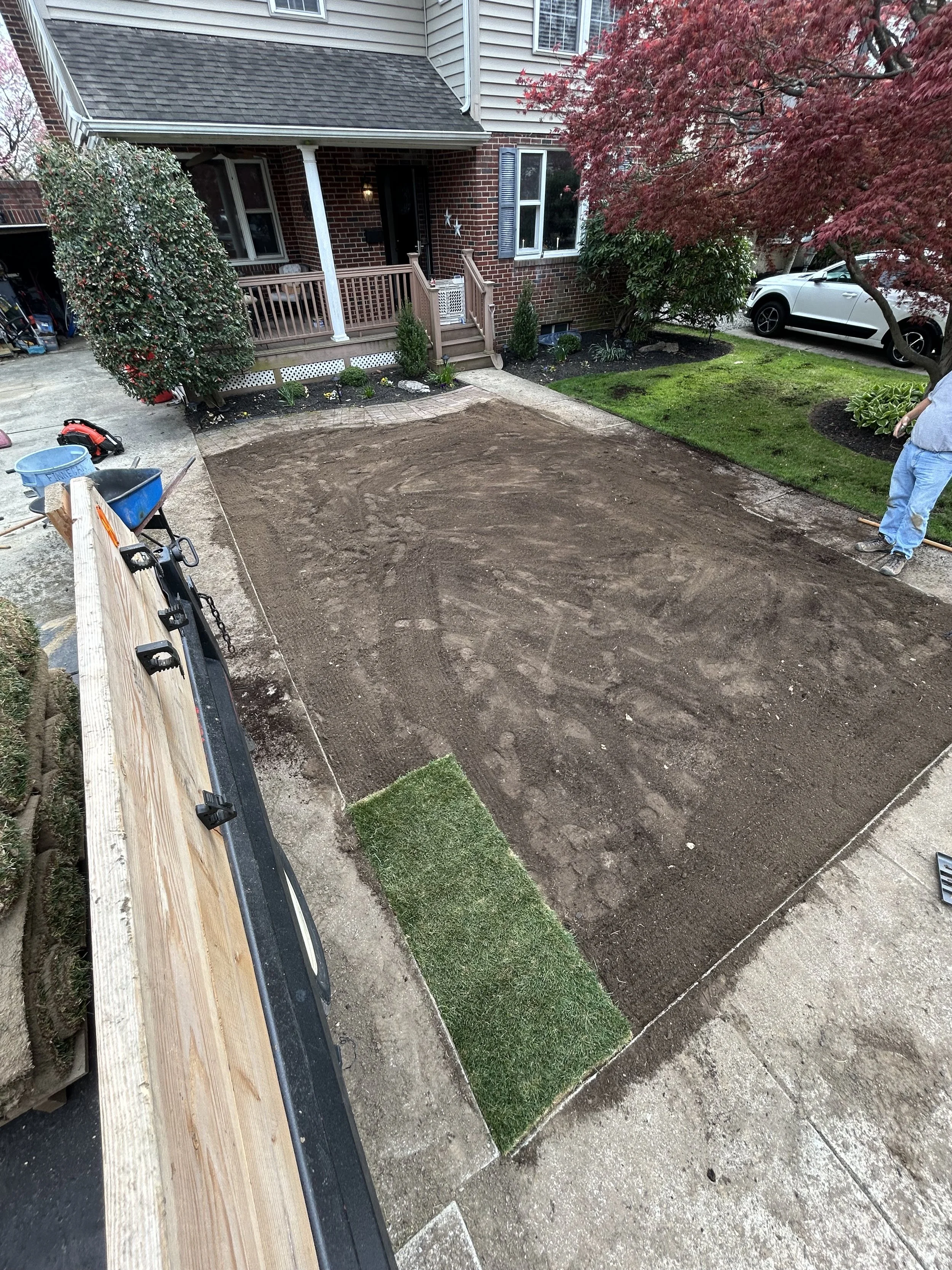 A backyard in front of a house with a partially completed lawn, showing a cleared dirt area, small patches of grass, and a person in work clothes holding a tool.