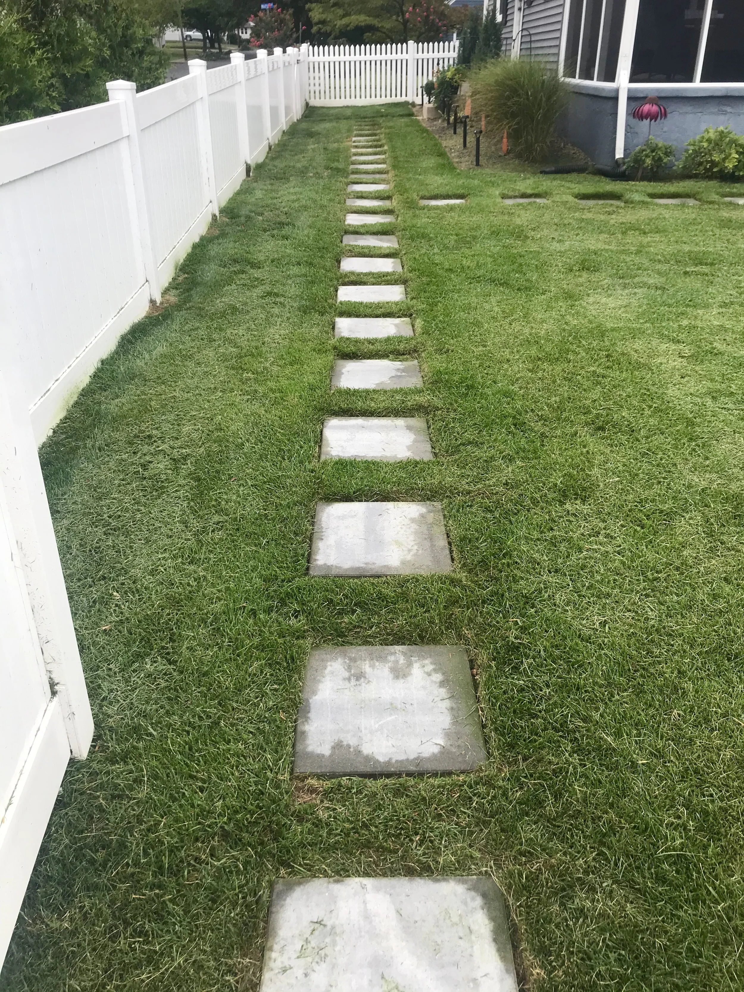 A straight row of concrete stepping stones on a grassy lawn, bordered by a white fence on the left and a house with plants on the right.