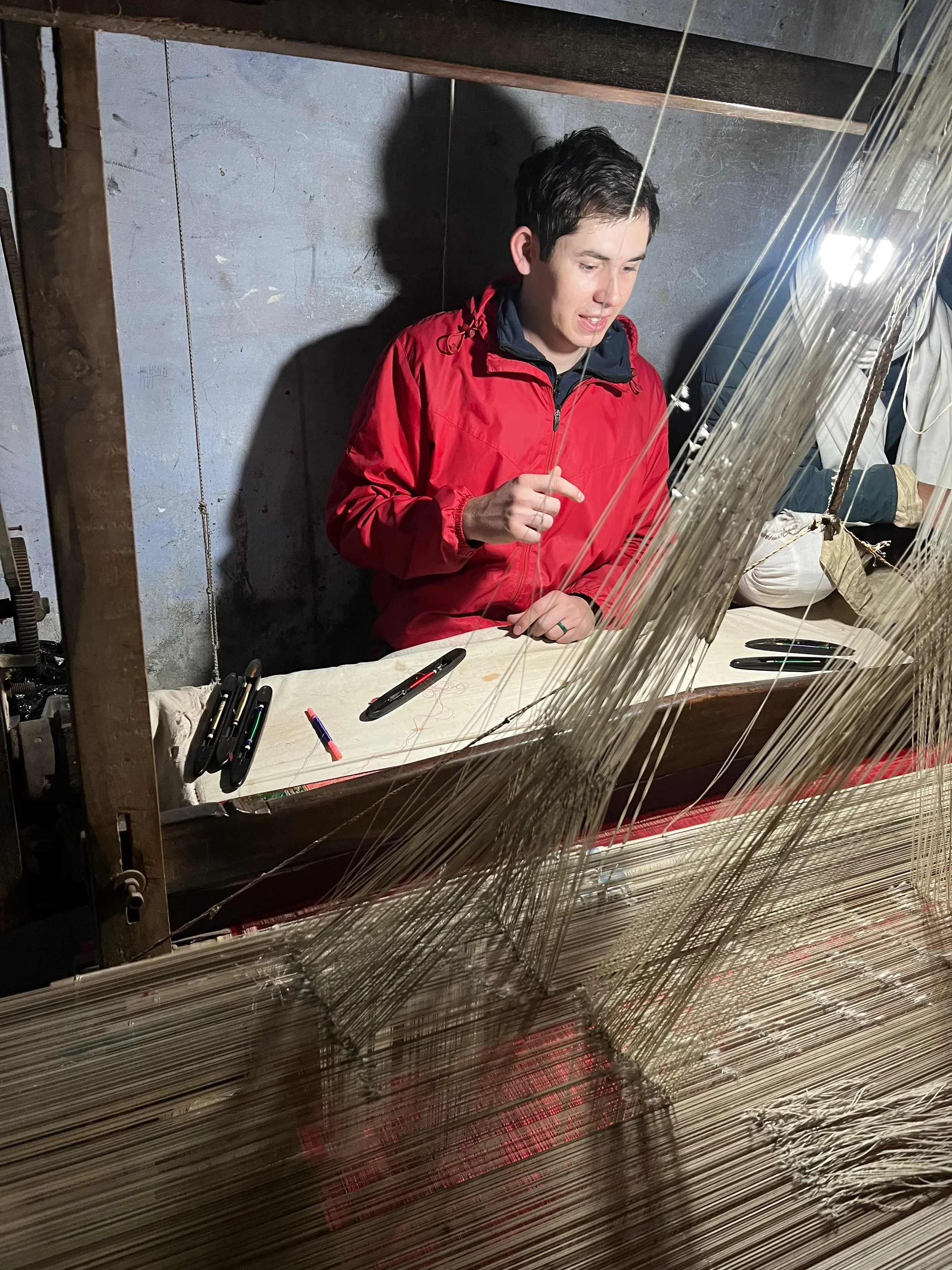 A young man wearing a red jacket sitting at a table with pens, surrounded by a weaving loom with numerous vertical threads, in an industrial or workshop setting.