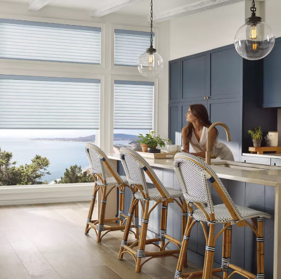 Woman sitting at kitchen island with blue cabinets, large windows, wicker chairs, and a view of water and trees outside.