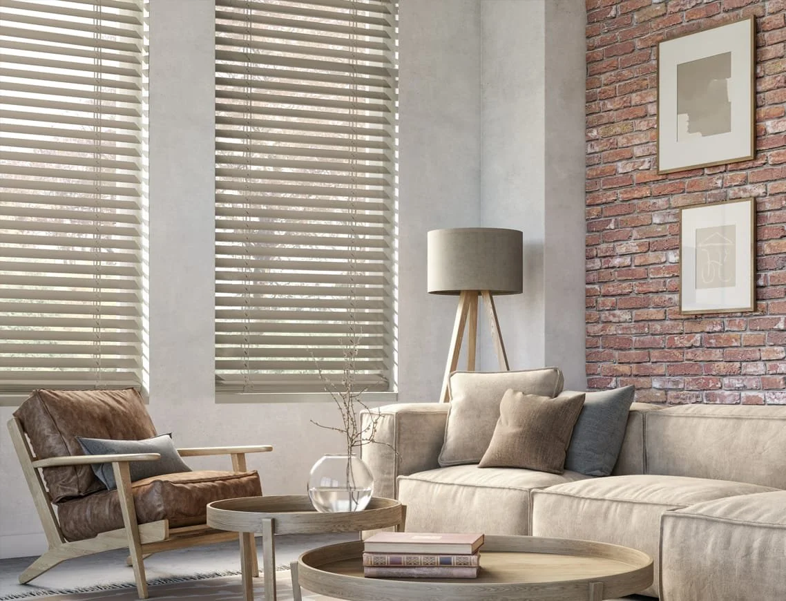 Living room with beige sectional sofa, wooden armchair with brown cushion, two round wooden coffee tables, a glass vase with twigs, and framed art on a red brick accent wall. Large windows with blinds letting in natural light, and a tripod floor lamp with a beige shade.
