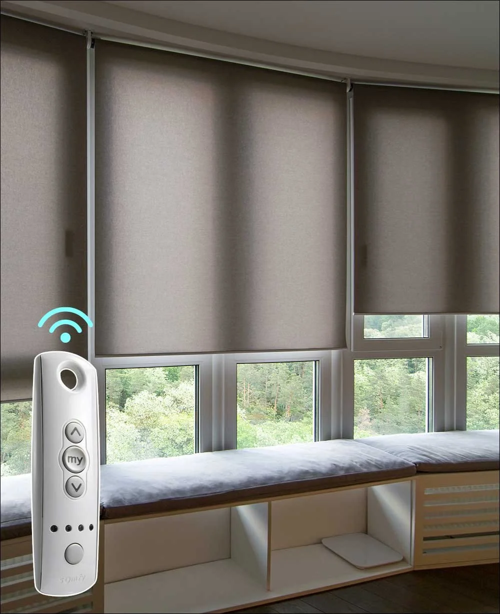 A room with large windows covered by beige roller blinds, a window seat with a cushion, and a view of trees outside. There's also a remote control with Wi-Fi symbol, likely for controlling the blinds.
