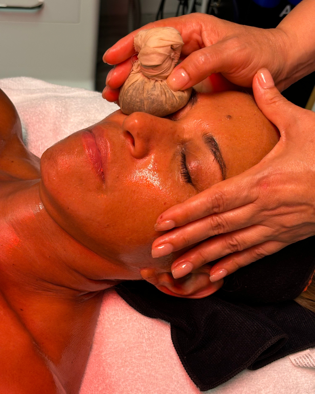 A woman receiving a facial massage with a herbal pinda on her eye, lying on a treatment bed with her eyes closed.