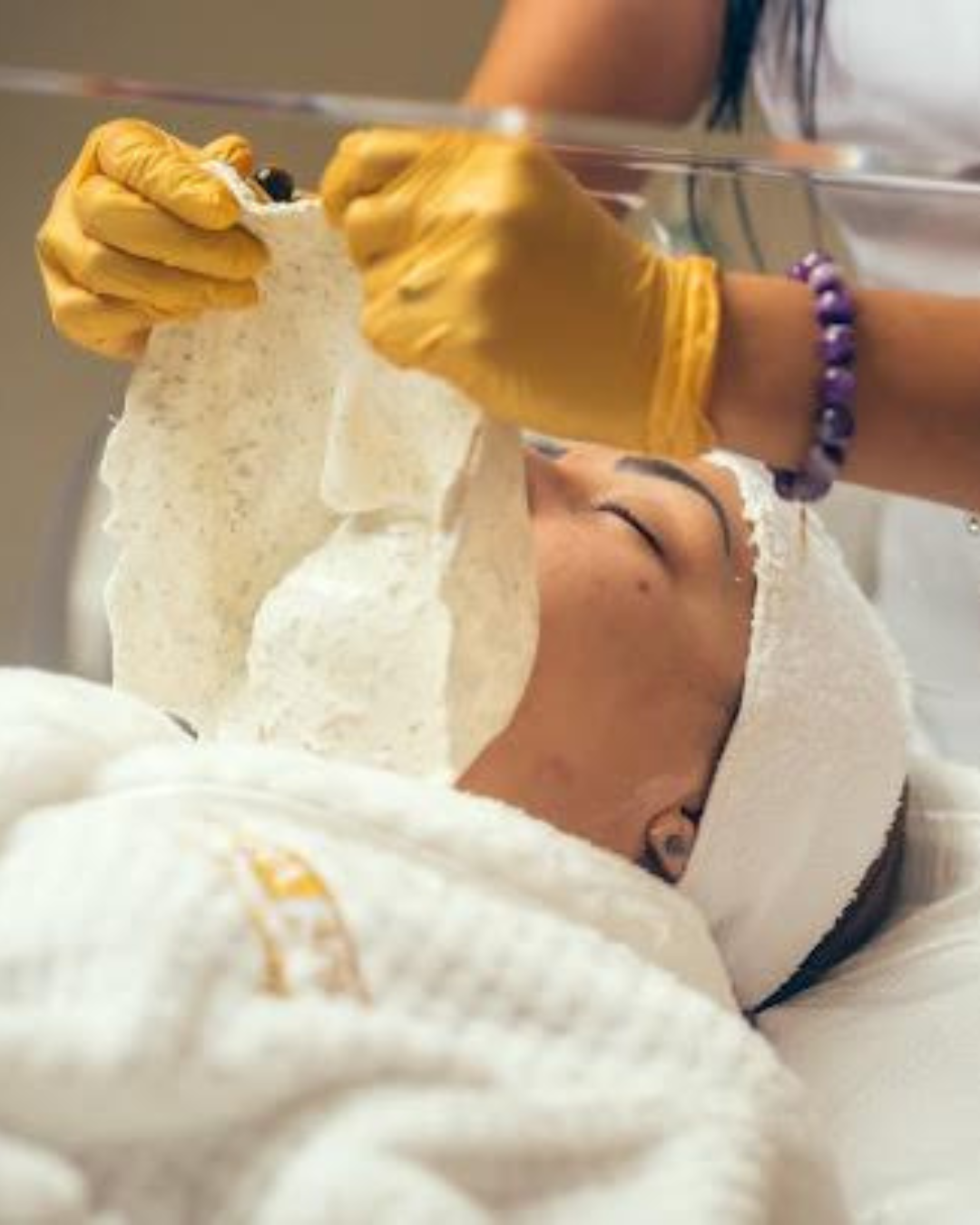 Technician wearing gold gloves removing a white facial mask from a client’s face.