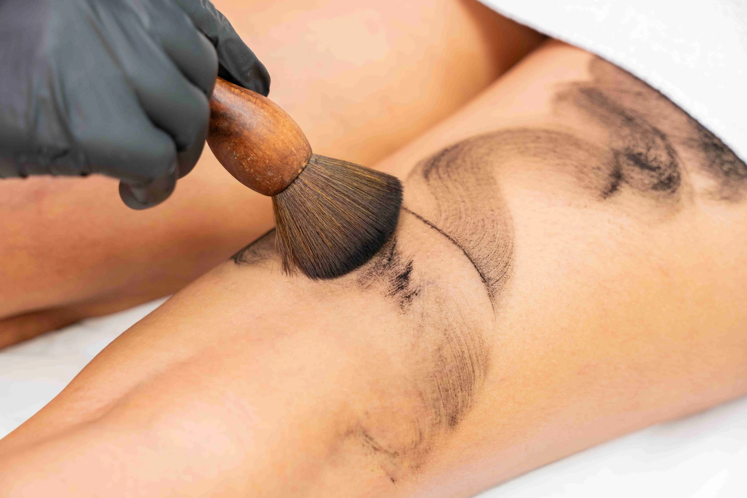 Technician wearing black gloves applying carbon powder treatment to a client’s legs