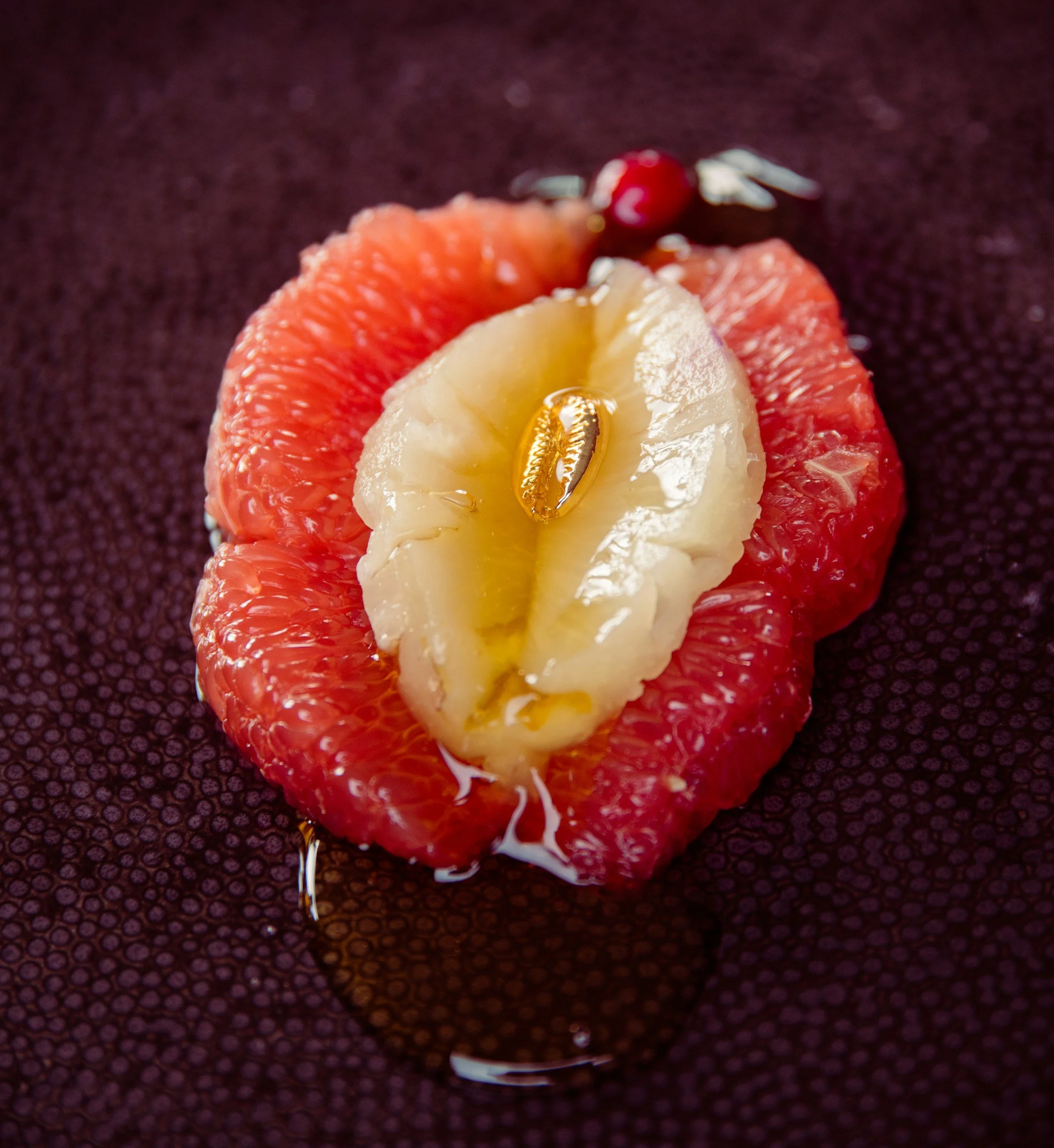 Close-up of a halved kumquat with a seed inside, placed on a textured dark surface with some liquid around it.