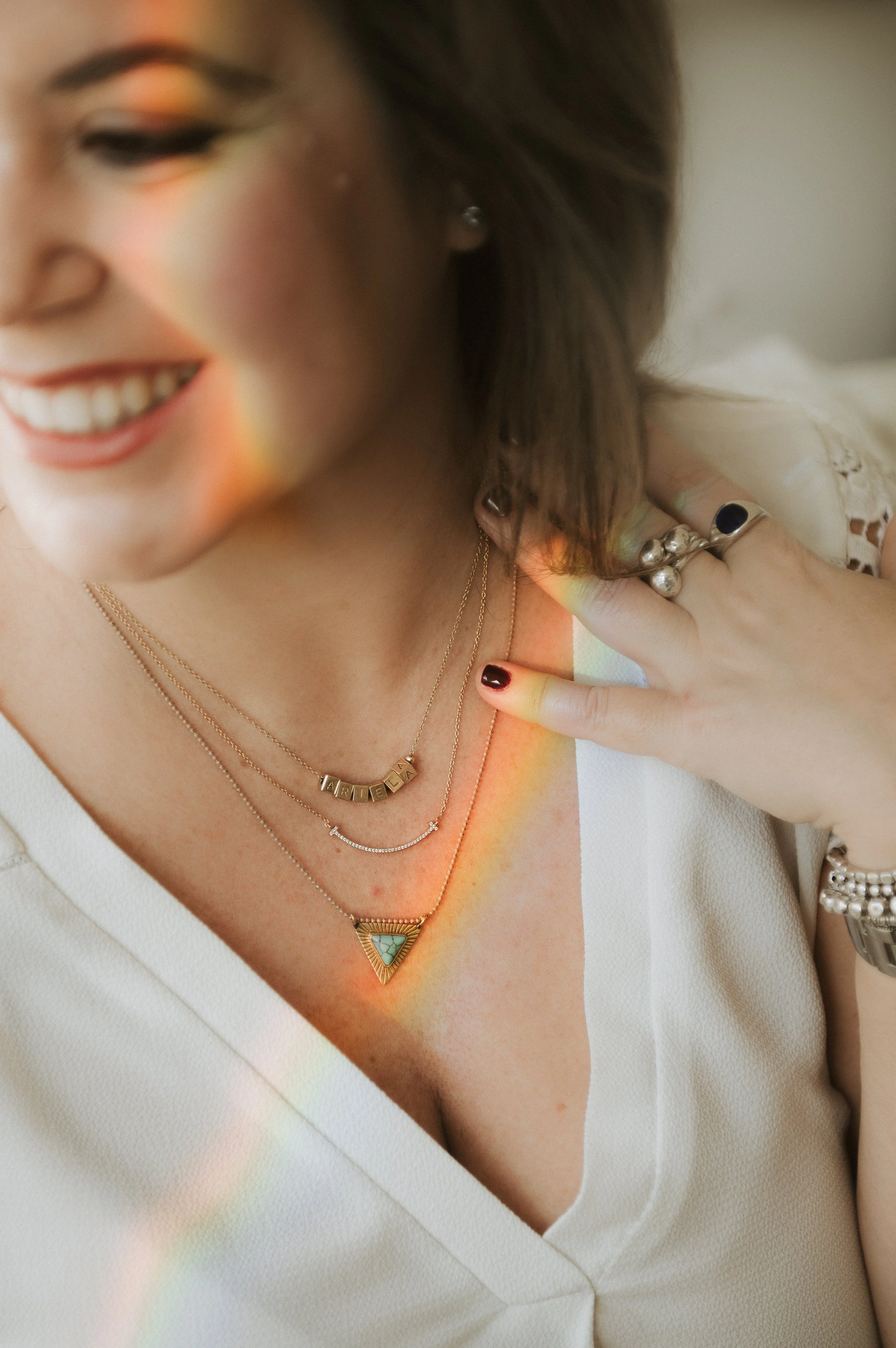 Close-up of a smiling woman wearing multiple layered necklaces, rings, and a watch, with rainbow light reflections on her skin and clothing.