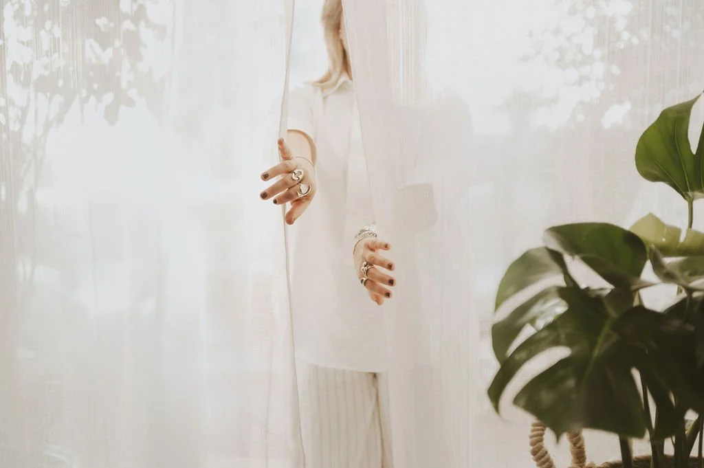 Person peeking from behind a white wall or curtain, wearing a sleeveless white top and striped pants, holding a curtain or fabric with her right hand, with jewelry on her fingers and wrist. There is a large leafy green plant on the right side of the image.