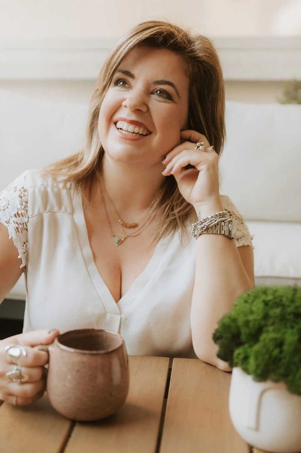A woman smiling, holding a coffee mug, sitting at a wooden table with a potted plant, wearing jewelry and a white top.