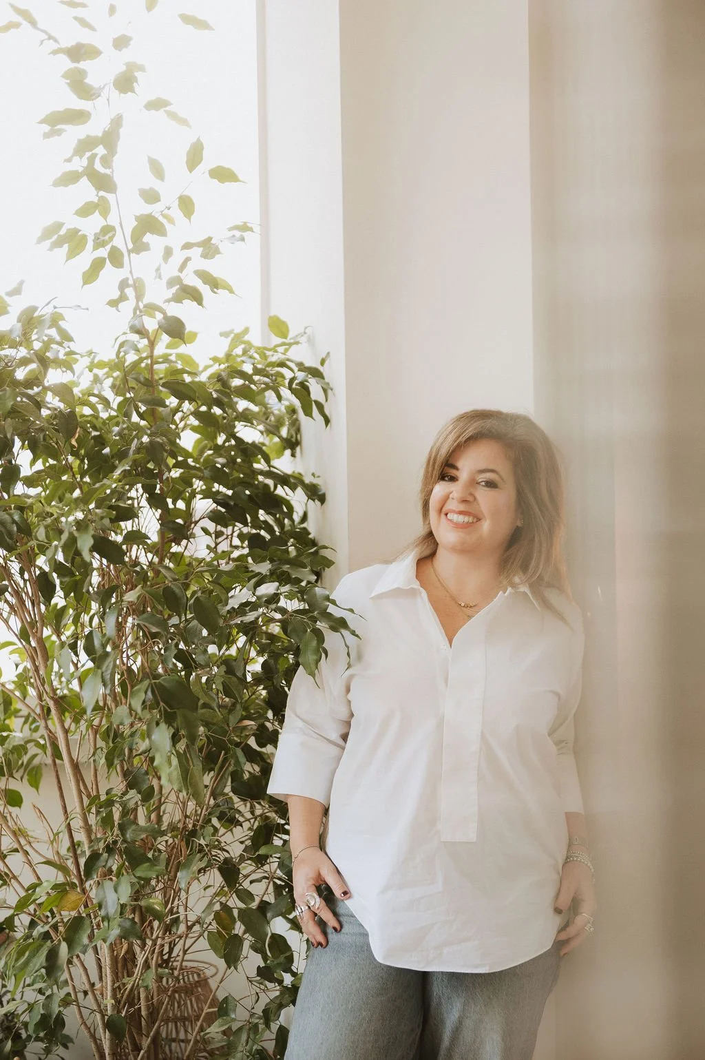 A woman with shoulder-length light brown hair, smiling, wearing a white blouse and gray pants, stands near a leafy green plant by a window with sheer curtains.