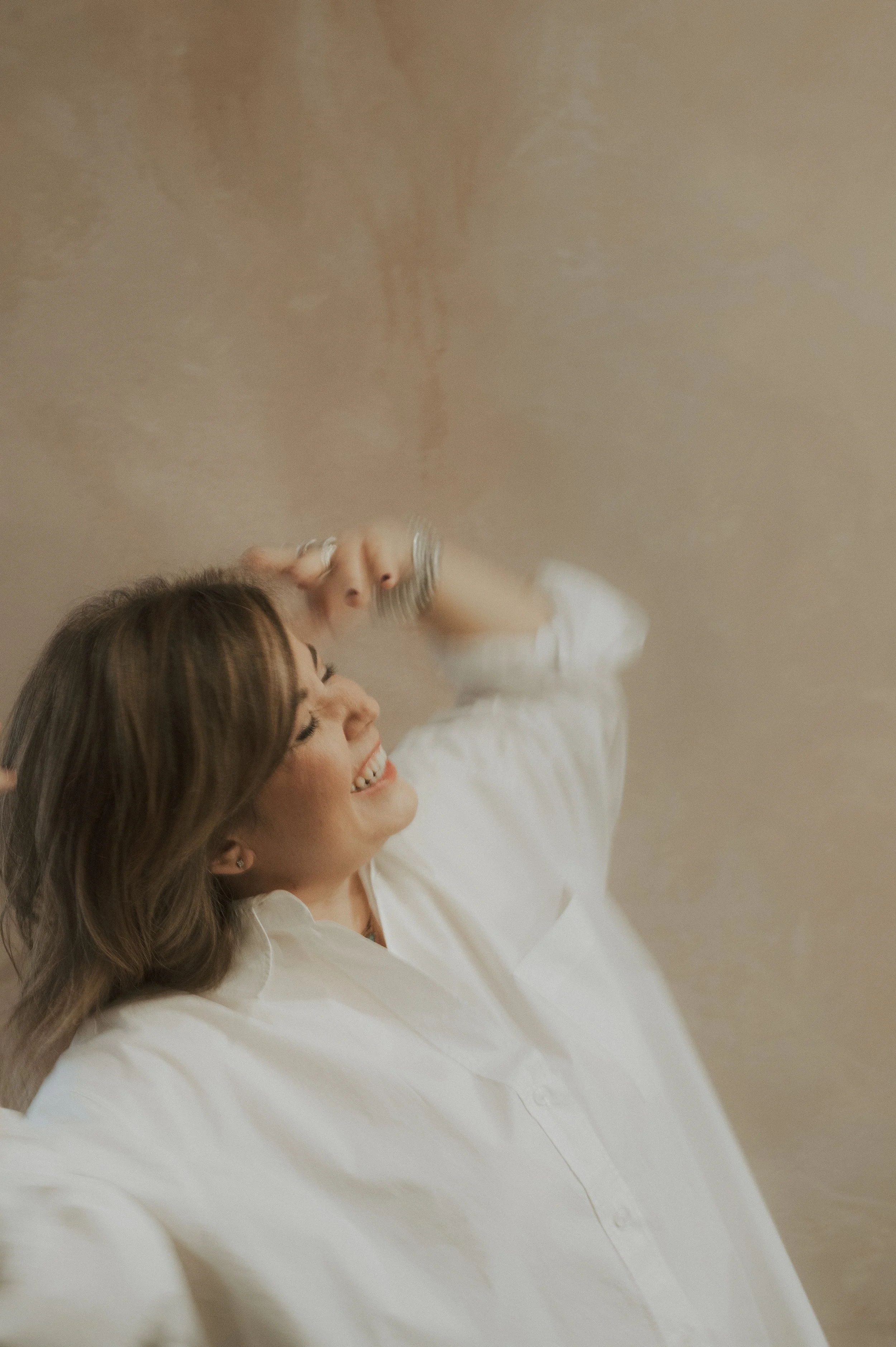 A woman with brown hair wearing a white shirt, smiling and laughing with her eyes closed, touching her head with one hand in front of a beige wall.