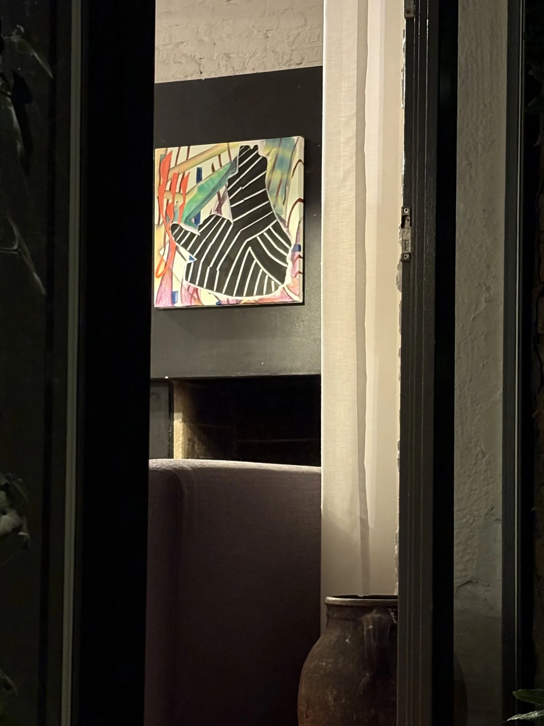 A framed colorful abstract painting on a black wall, with a large pottery vase on a table in the foreground, and a window with dark frames.