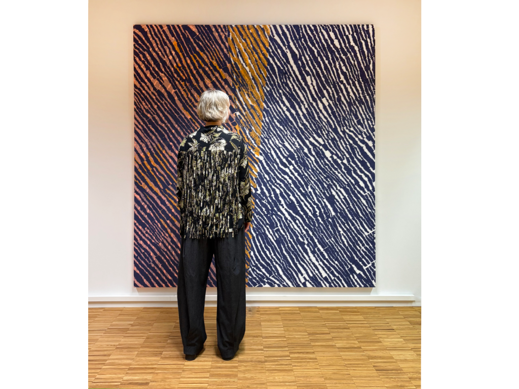A person with gray hair wearing a black and white patterned jacket looks at an abstract painting with diagonal orange, black, white, and blue lines in an art gallery with light-colored walls and wooden flooring.