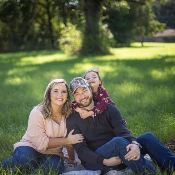 Family of three sitting on grass in a park, smiling and enjoying a sunny day. The woman is wearing a light pink sweater, the man is wearing a black hoodie, and the girl is wearing a maroon sweater and hugging the man from behind.