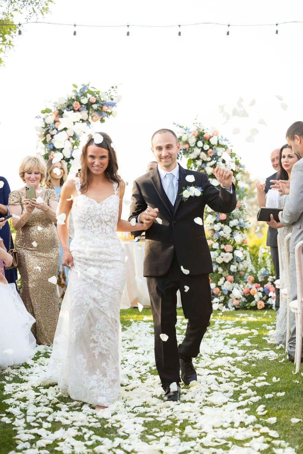 A bride and groom walking down the aisle, smiling, with rose petal confetti falling at an outdoor wedding ceremony decorated with floral arrangements.