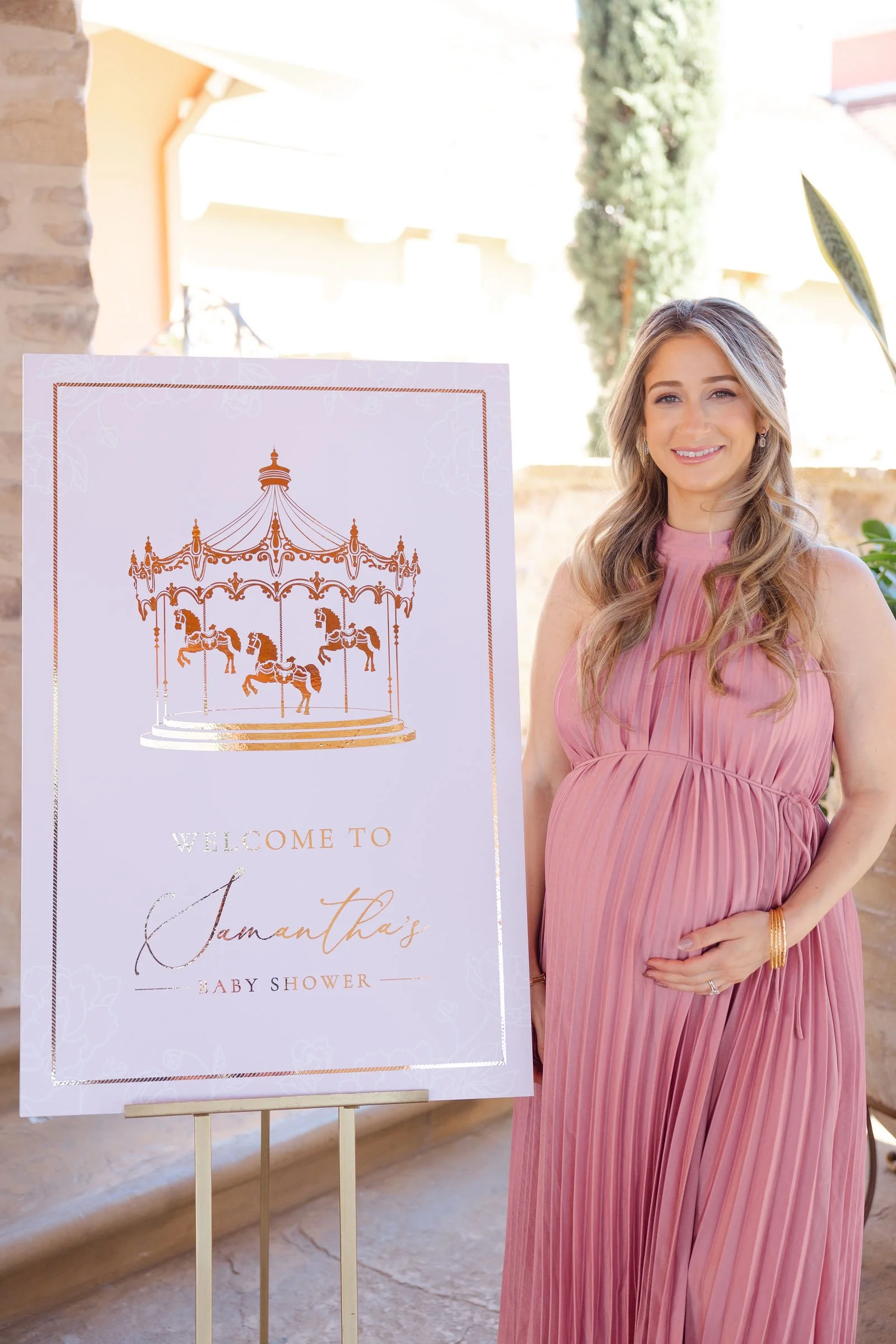 Pregnant woman in pink dress standing next to a sign welcoming guests to a baby shower.