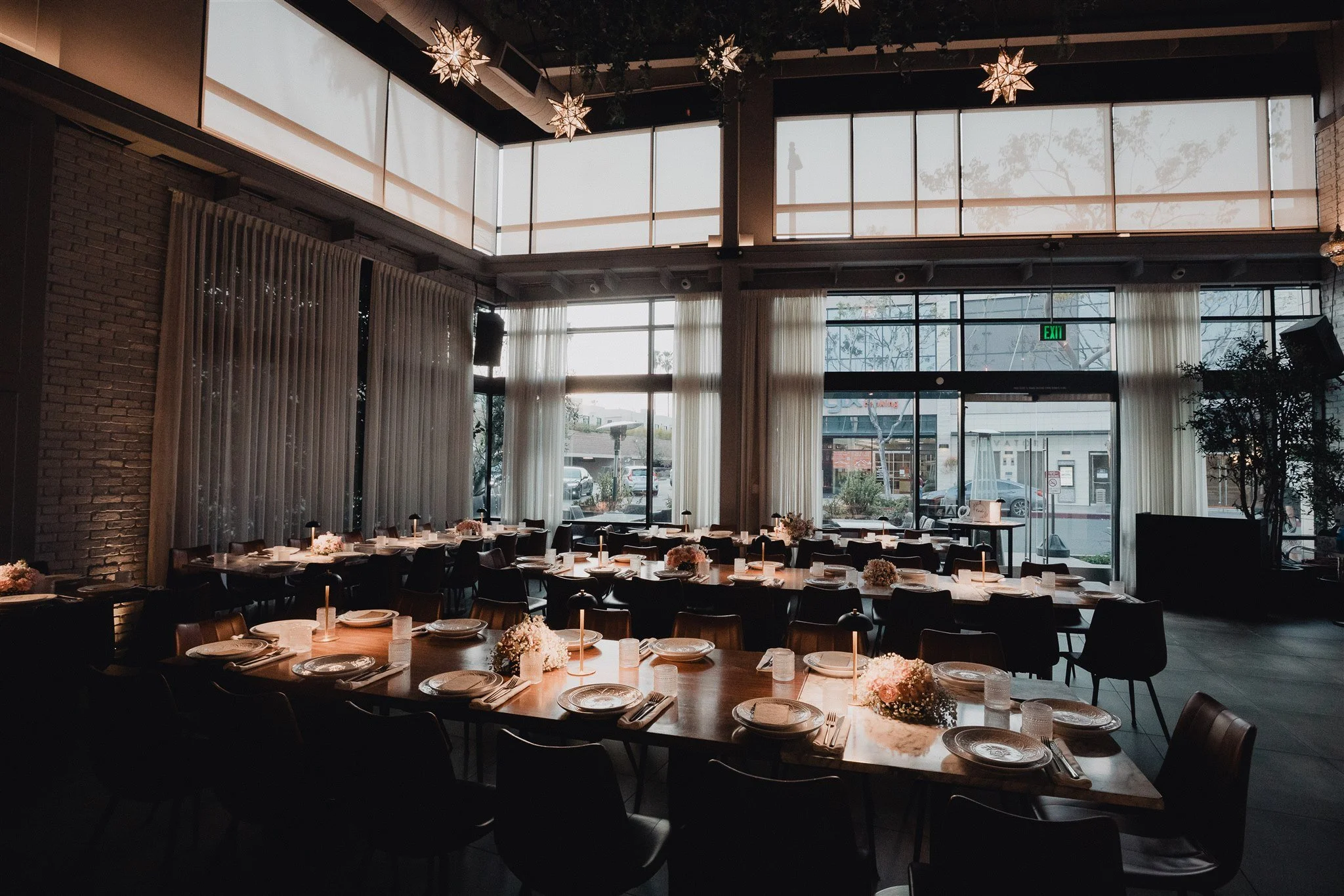 Interior of a restaurant or event space with long tables set for a meal, decorated with flowers and candles, featuring large windows with white curtains and star-shaped hanging lights.