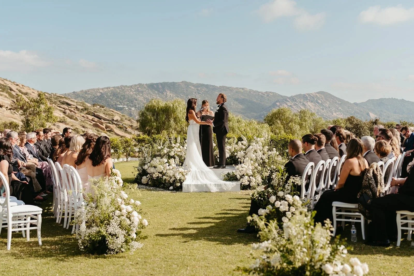 Outdoor wedding ceremony with a bride and groom exchanging vows on a platform decorated with white flowers, surrounded by seated guests in a scenic mountain landscape on a sunny day.