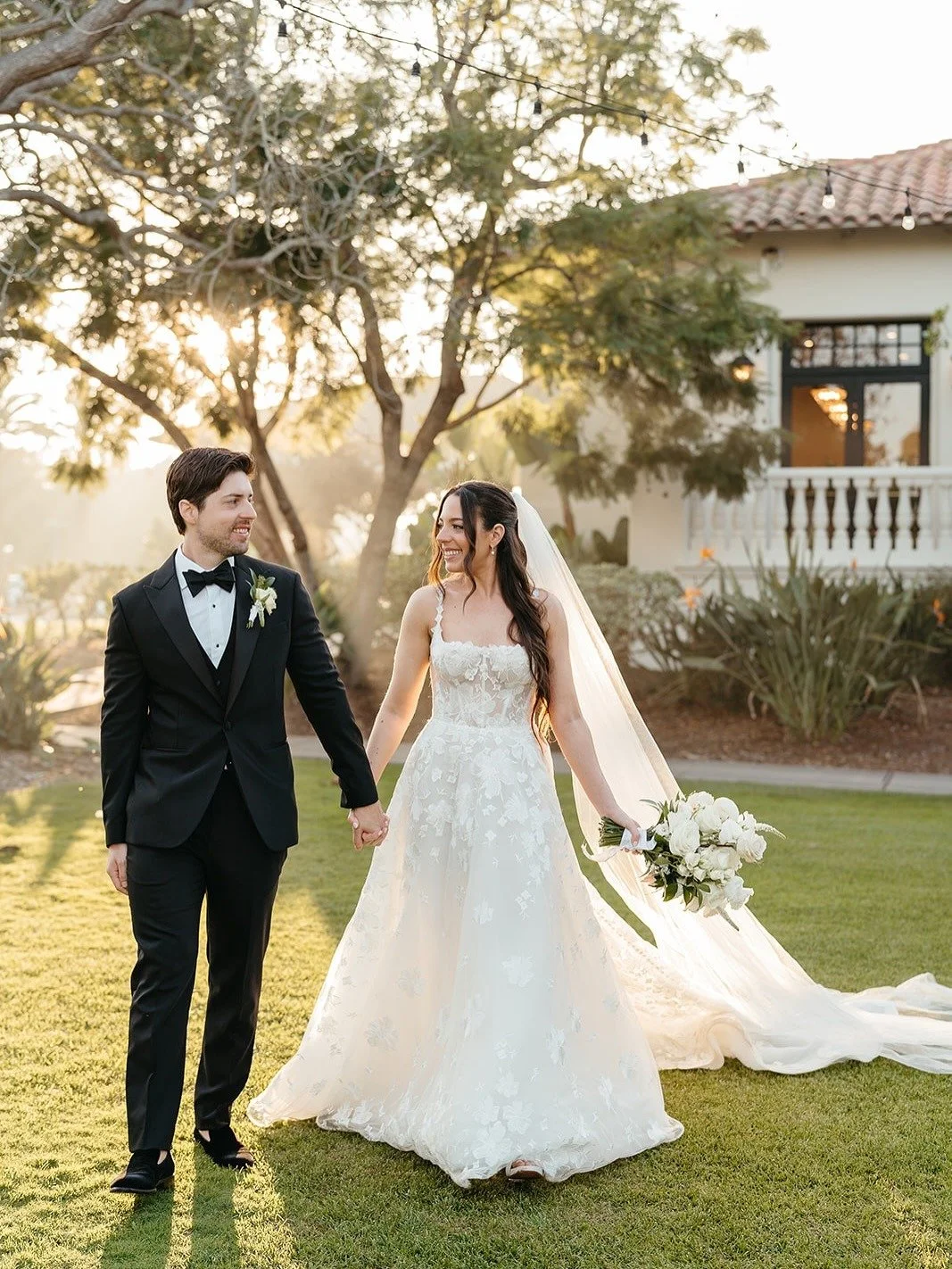 A newlywed couple walking hand in hand outdoors during sunset, the bride holding a bouquet of white flowers and wearing a lace wedding gown with a long train, the groom in a black tuxedo, with a house and trees in the background.
