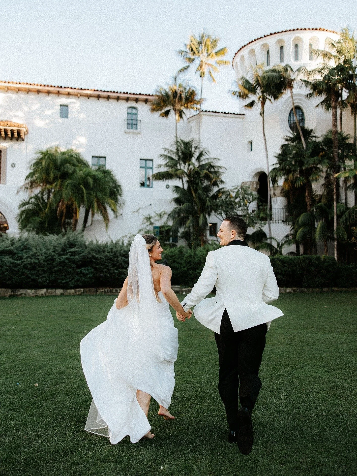 A bride and groom holding hands and running on a grassy lawn with a white Mediterranean-style building and tall palm trees in the background.