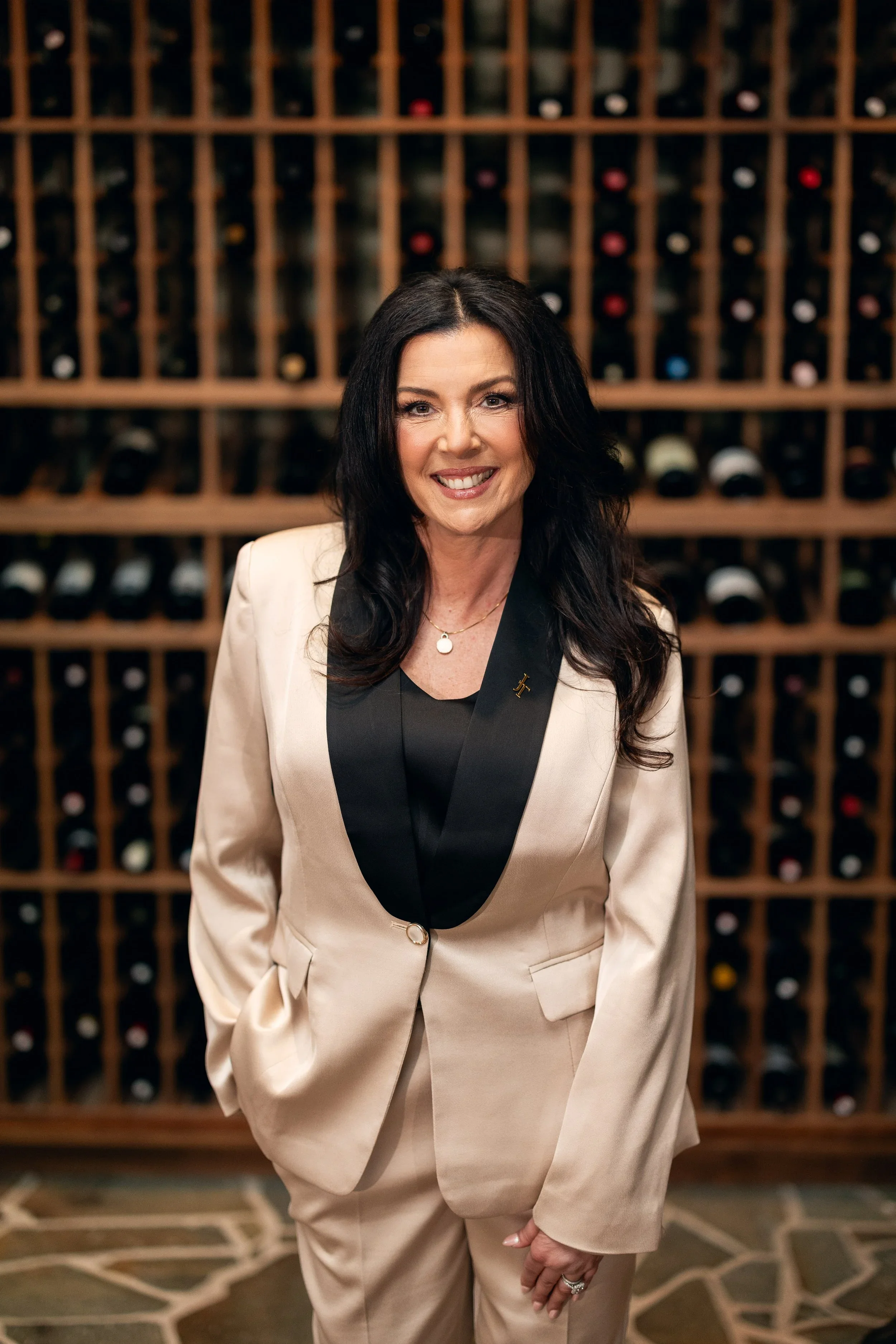 A woman with long dark hair smiling in front of a wine rack, wearing a cream suit with black lapels and a black shirt.