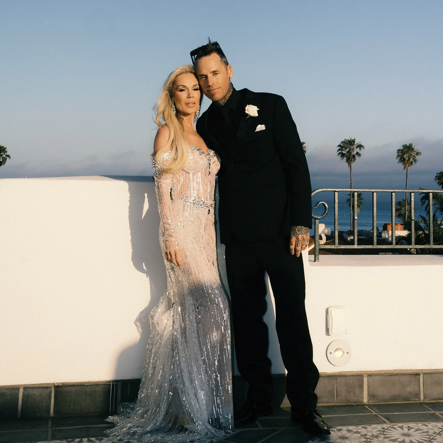 A woman in a shimmering, embellished gown stands next to a man in a black suit on a balcony with a view of palm trees and the ocean in the background.