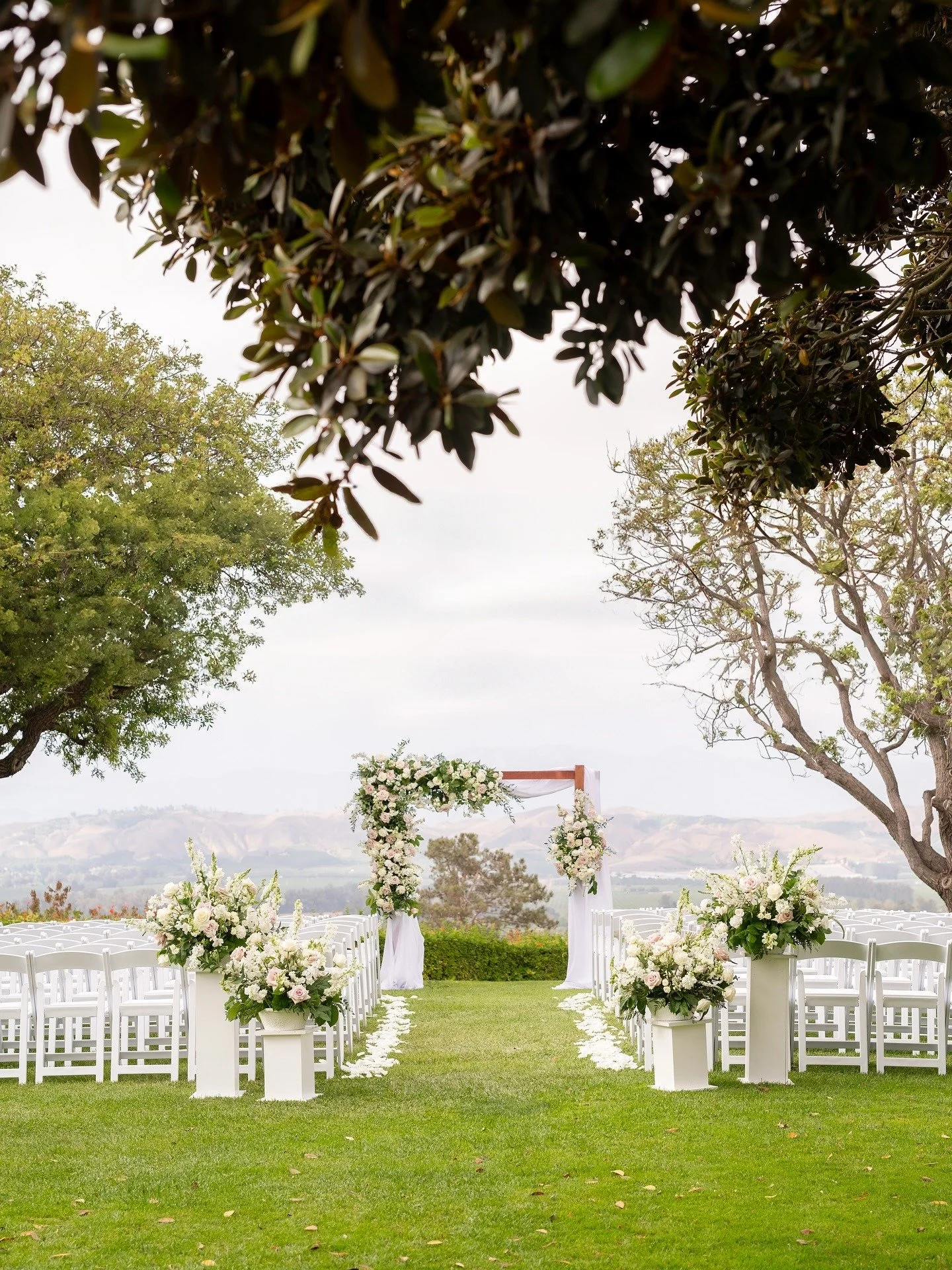 Outdoor wedding ceremony setup with white chairs, floral arrangements, and an arch with flowers, on a green lawn with trees and distant hills in the background.