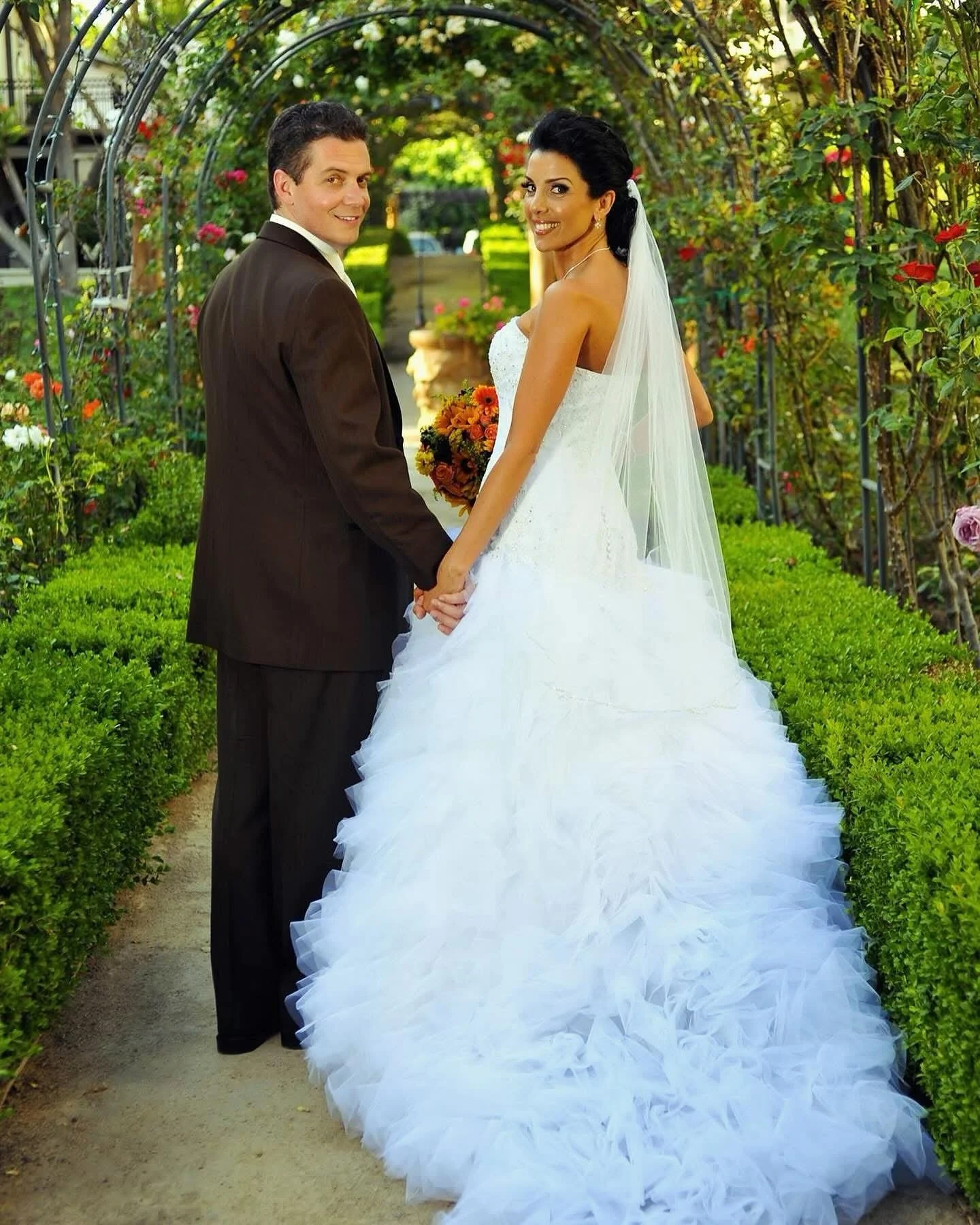 A bride and groom holding hands in a garden archway with greenery and flowers, the bride in a white wedding gown with a veil, the groom in a dark suit, both smiling at the camera.
