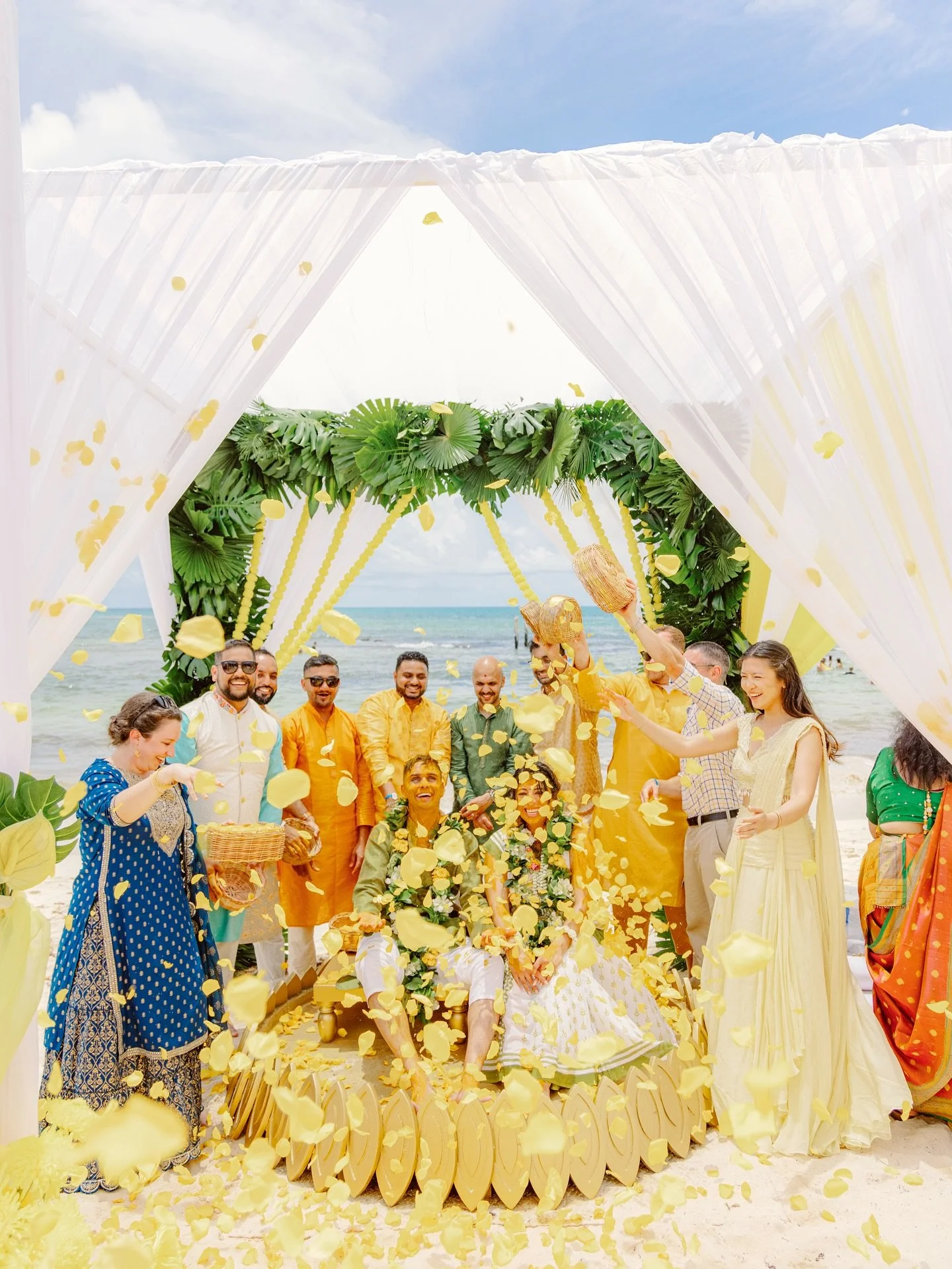 A wedding celebration on a beach with a large group of people, a decorated canopy with green leaves, and yellow flowers, with yellow flower petals falling and a sunny sky.