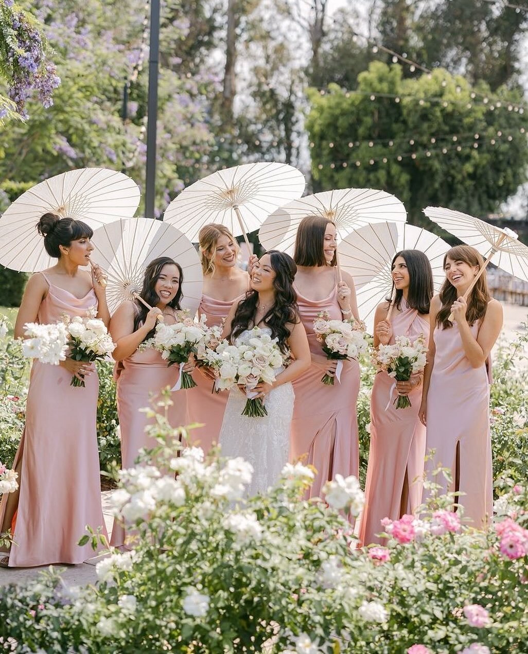 A bride and six bridesmaids in pink dresses standing outdoors with white parasols and bouquets of white flowers, surrounded by flowering bushes and trees.