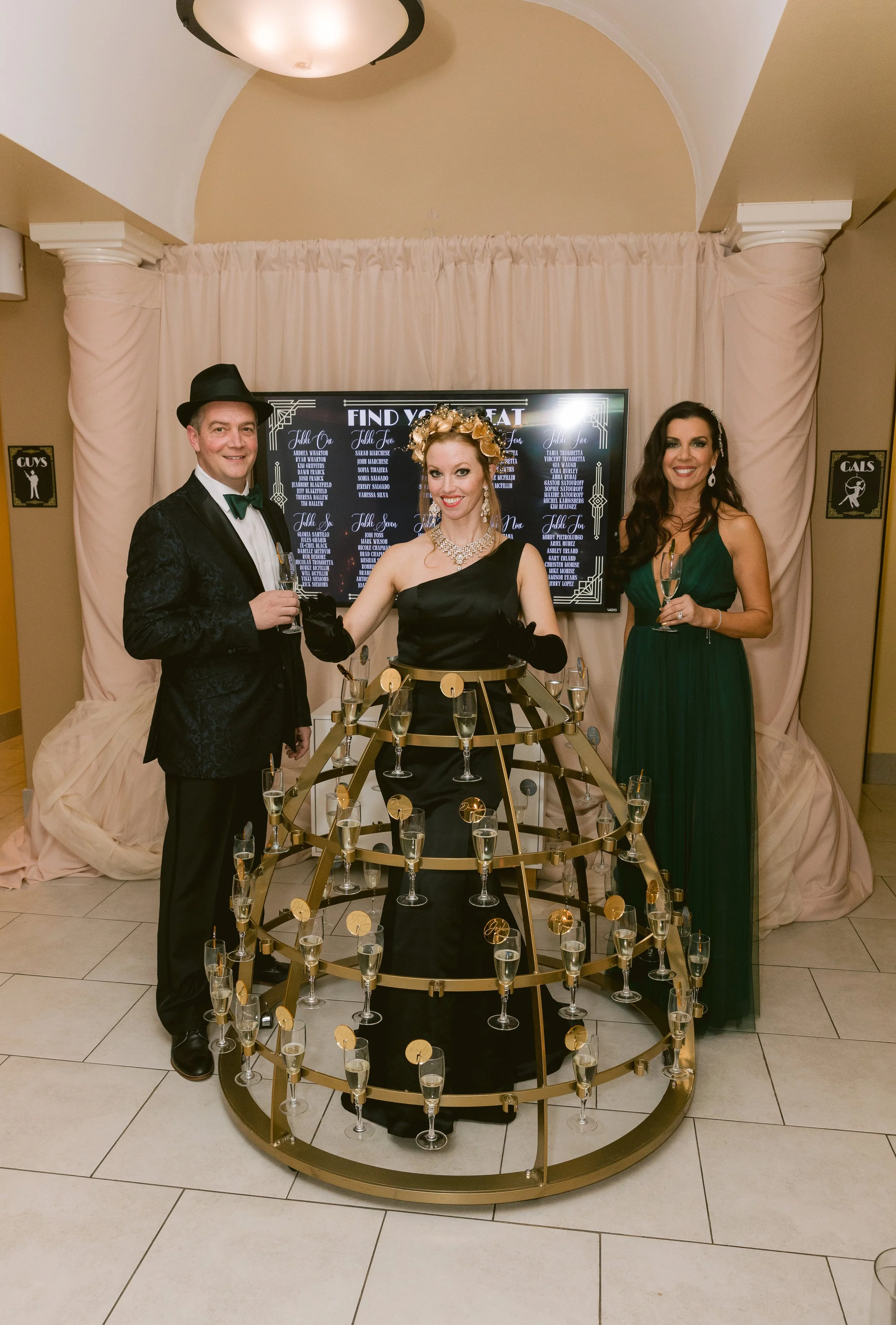 Three people dressed in formal attire celebrating at a party. They are standing behind a tiered display stand with glasses of champagne on it. The man on the left is wearing a black suit with a bow tie and a hat. The woman in the middle is wearing a 