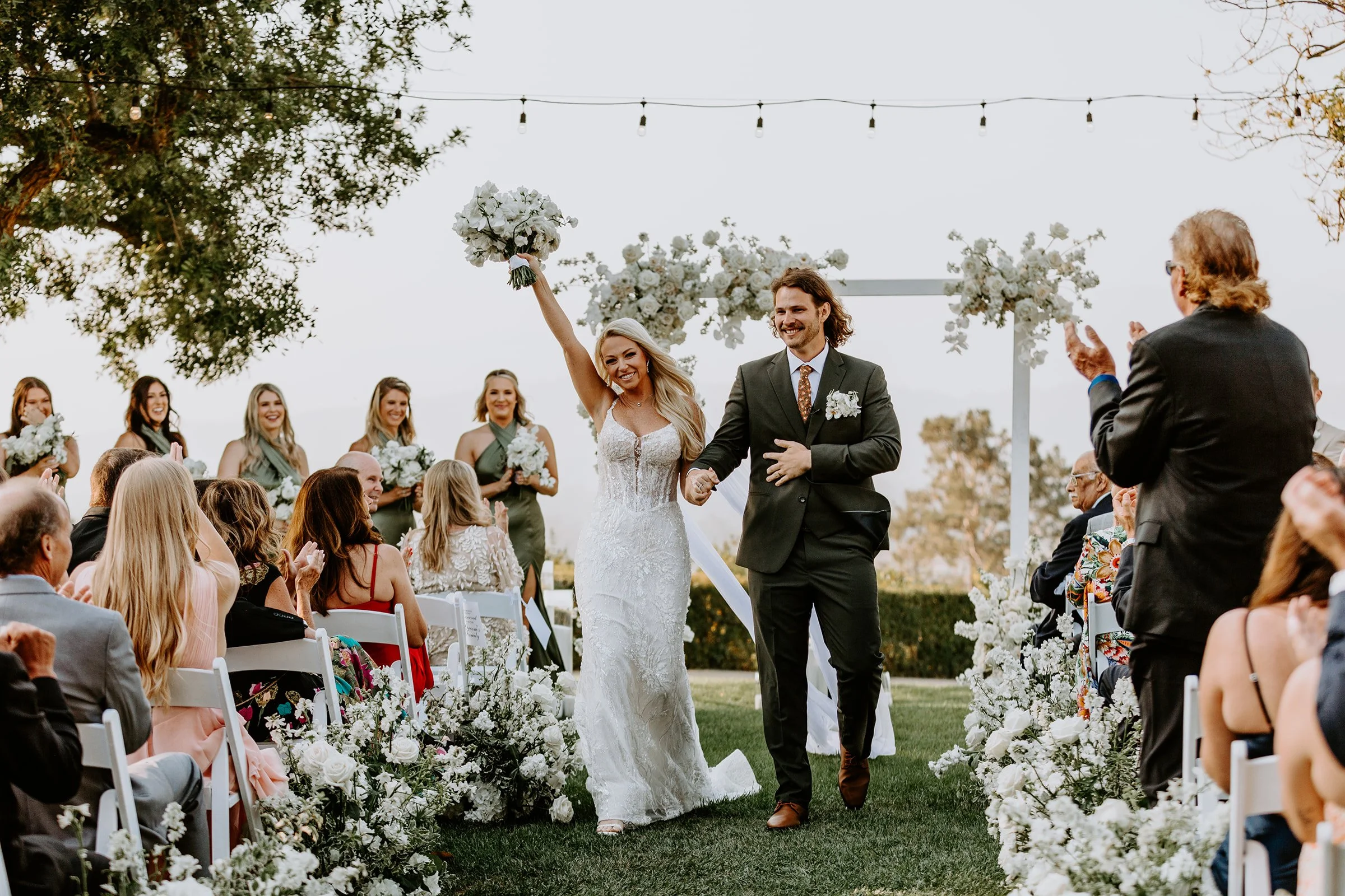 A bride and groom walking down the aisle at an outdoor wedding ceremony, with guests seated on either side and bridesmaids in green dresses in the background. The bride is holding a bouquet and raising her hand, smiling, while the groom is holding her hand and smiling.