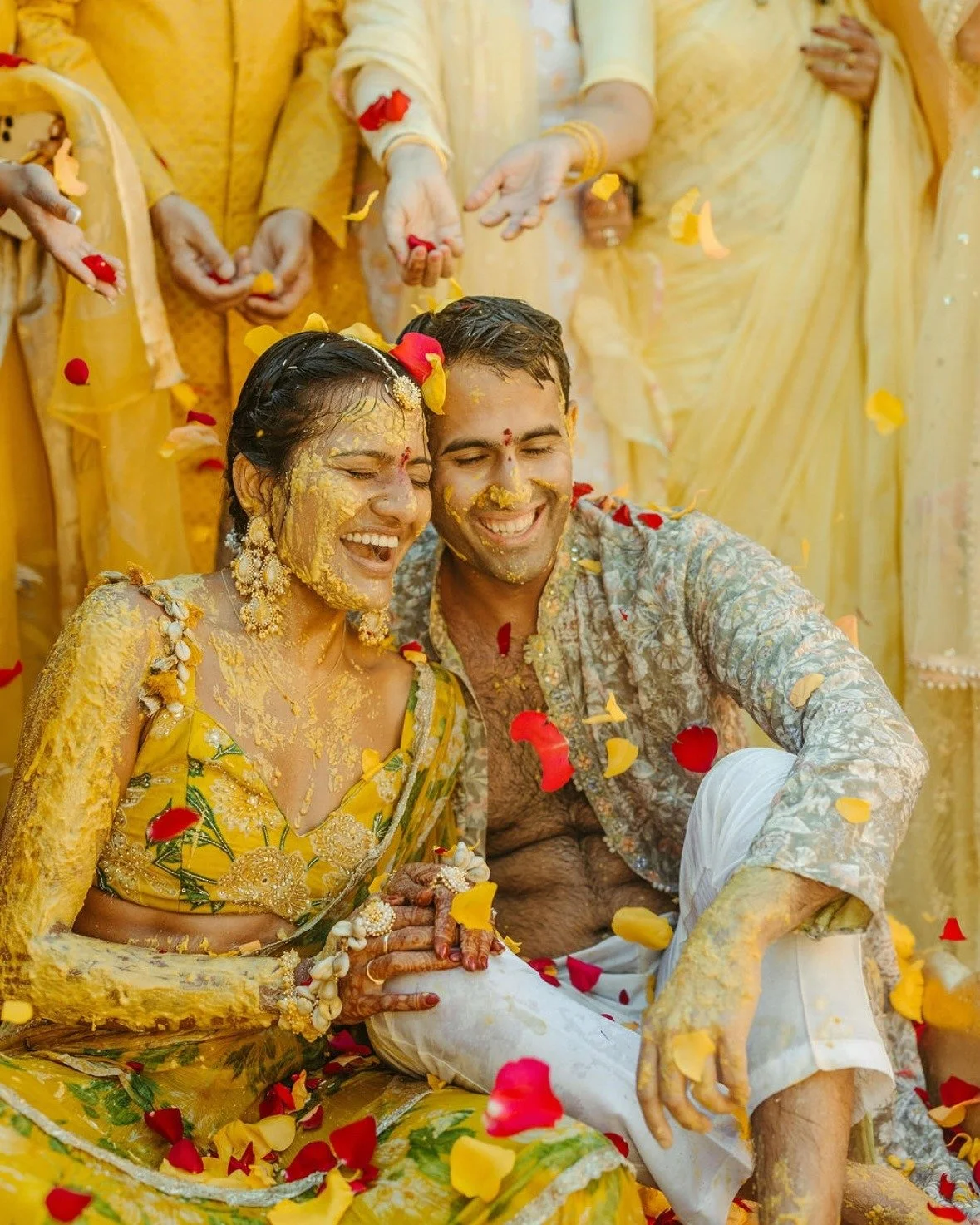 A newlywed couple celebrates their Indian wedding in vibrant yellow attire, covered in turmeric paste, surrounded by flower petals, during a joyful ceremony.