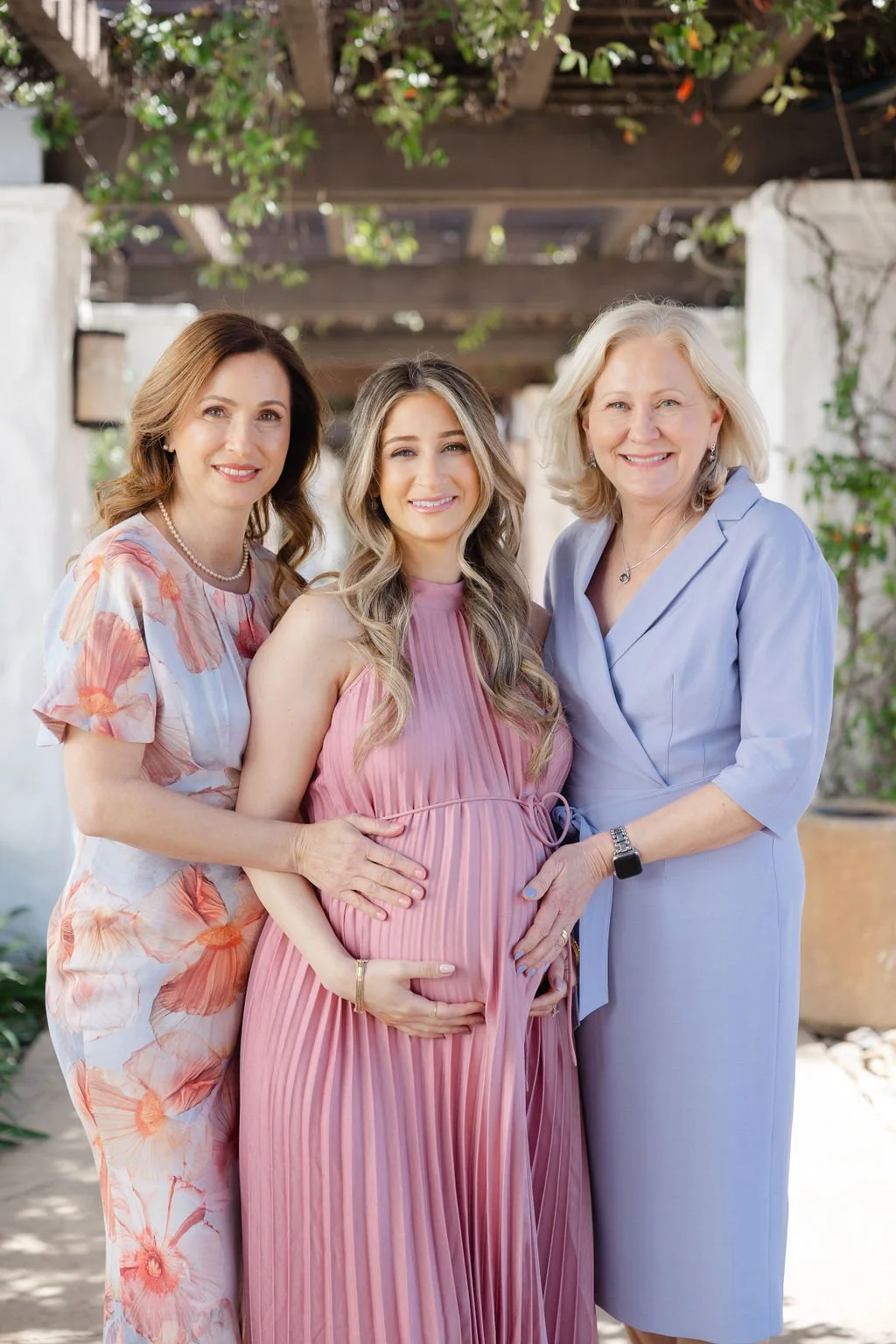 Three women, one pregnant, standing outdoors under a wooden trellis with greenery, smiling at the camera.