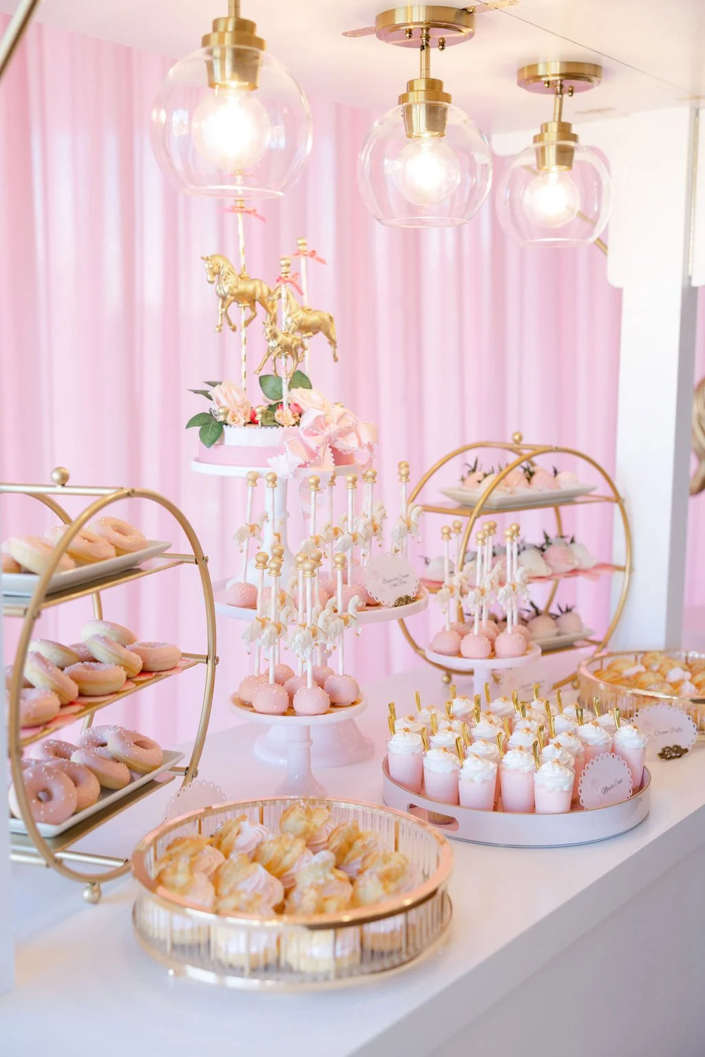 A dessert table decorated with pink and gold accents, featuring donuts, cake pops, and cookies, with a pink backdrop and elegant lighting.