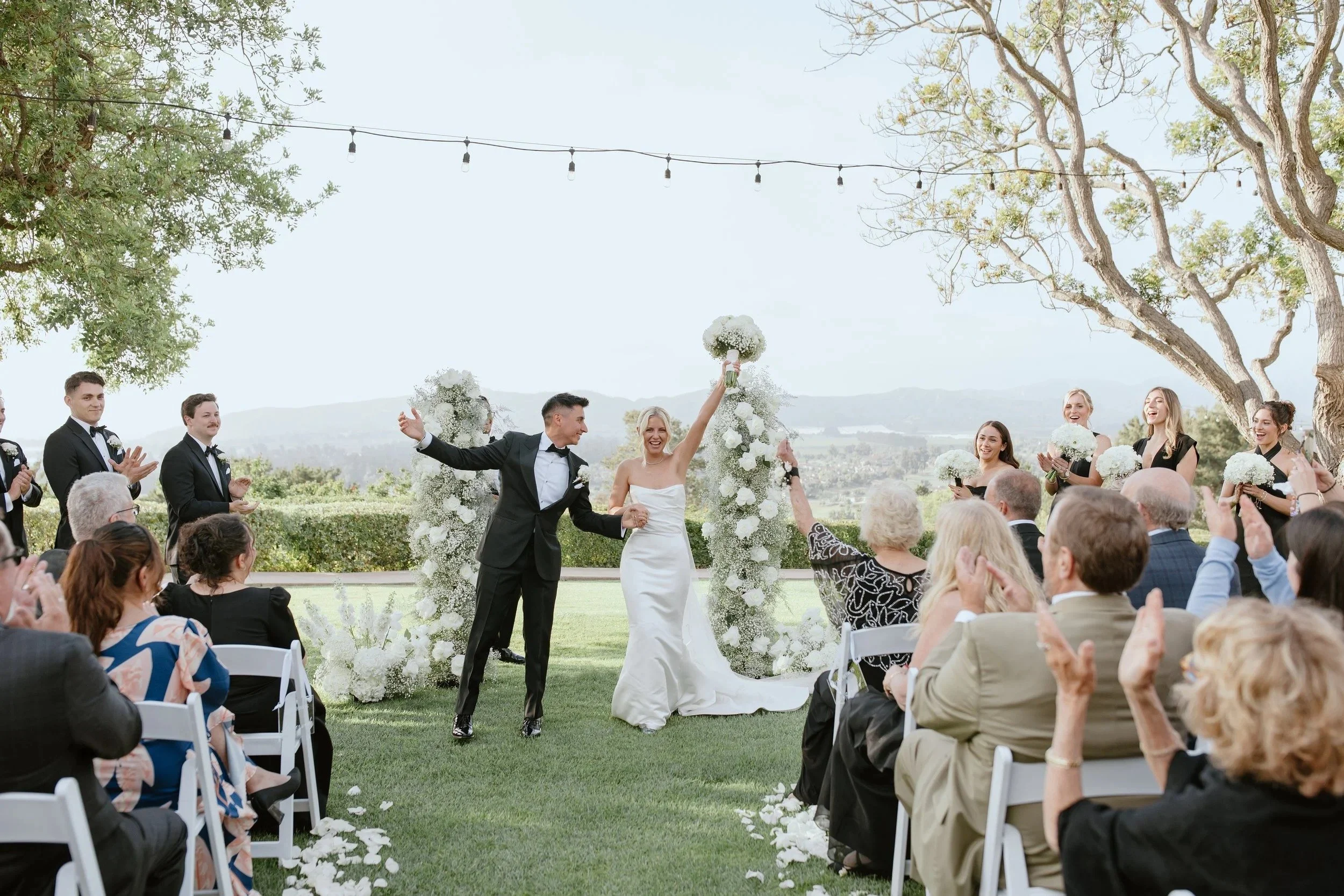 A wedding ceremony outdoors with the bride and groom holding hands, surrounded by friends and family, with decorative floral arrangements and string lights overhead.
