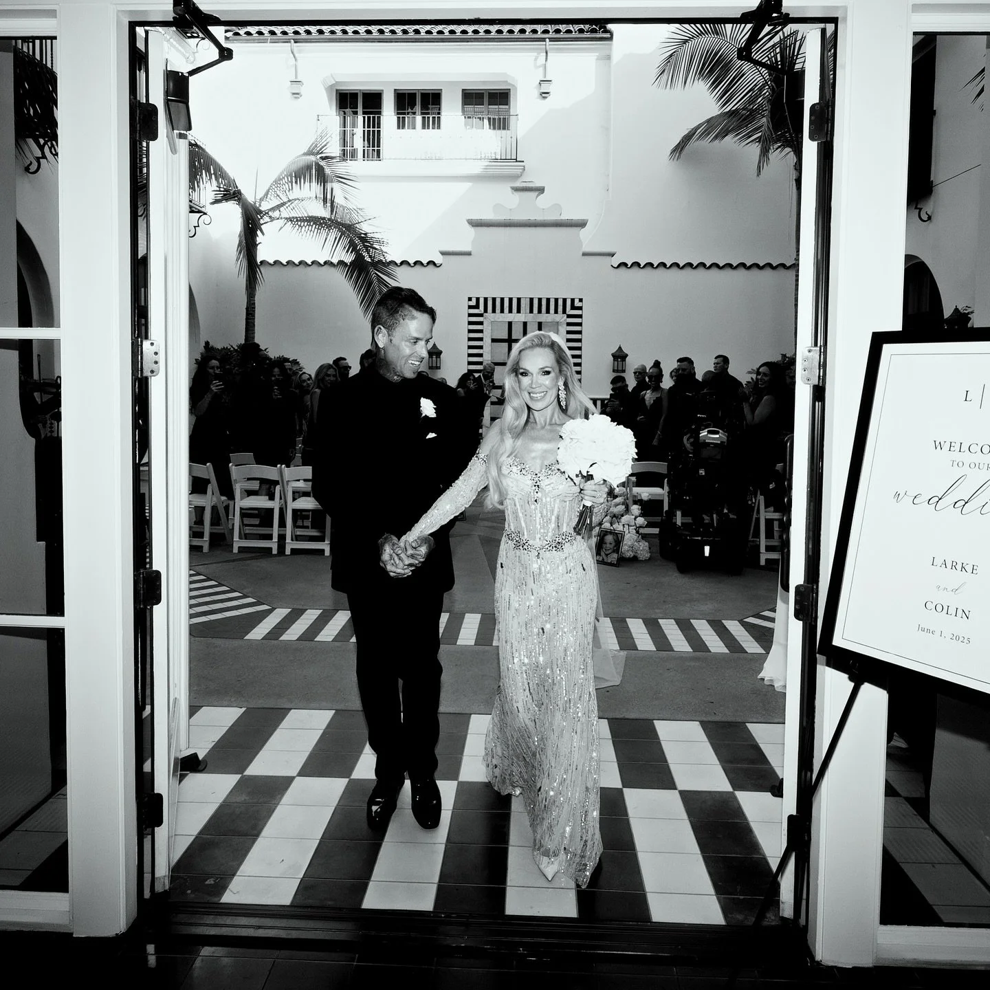 A bride and groom walk hand in hand through a doorway into their wedding reception, smiling. The bride is holding a bouquet of flowers, and the groom is dressed in a dark tuxedo. Guests are seated in the background, and a welcoming sign with wedding 