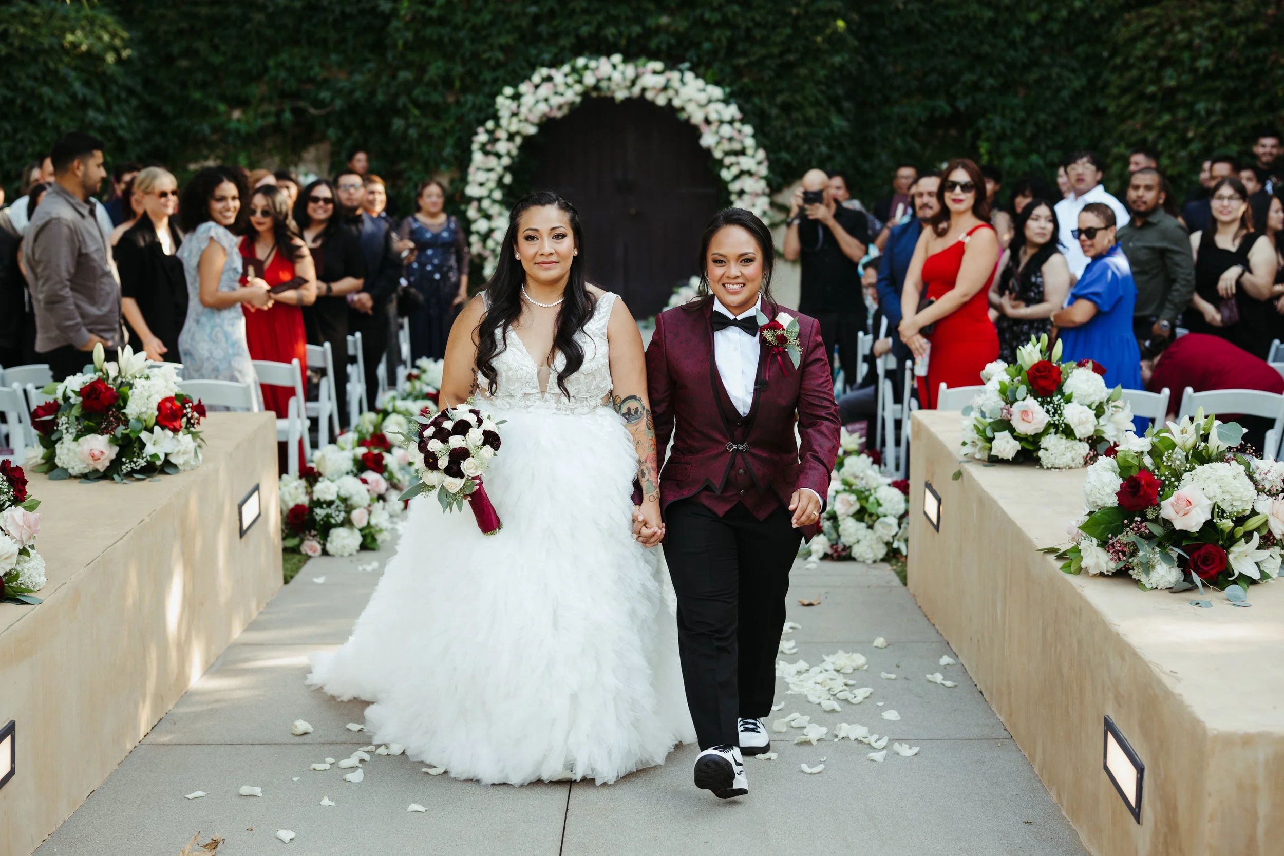 Two brides holding hands, one in a white wedding dress and the other in a red blazer, walking down the aisle at a wedding ceremony surrounded by guests and floral decorations.