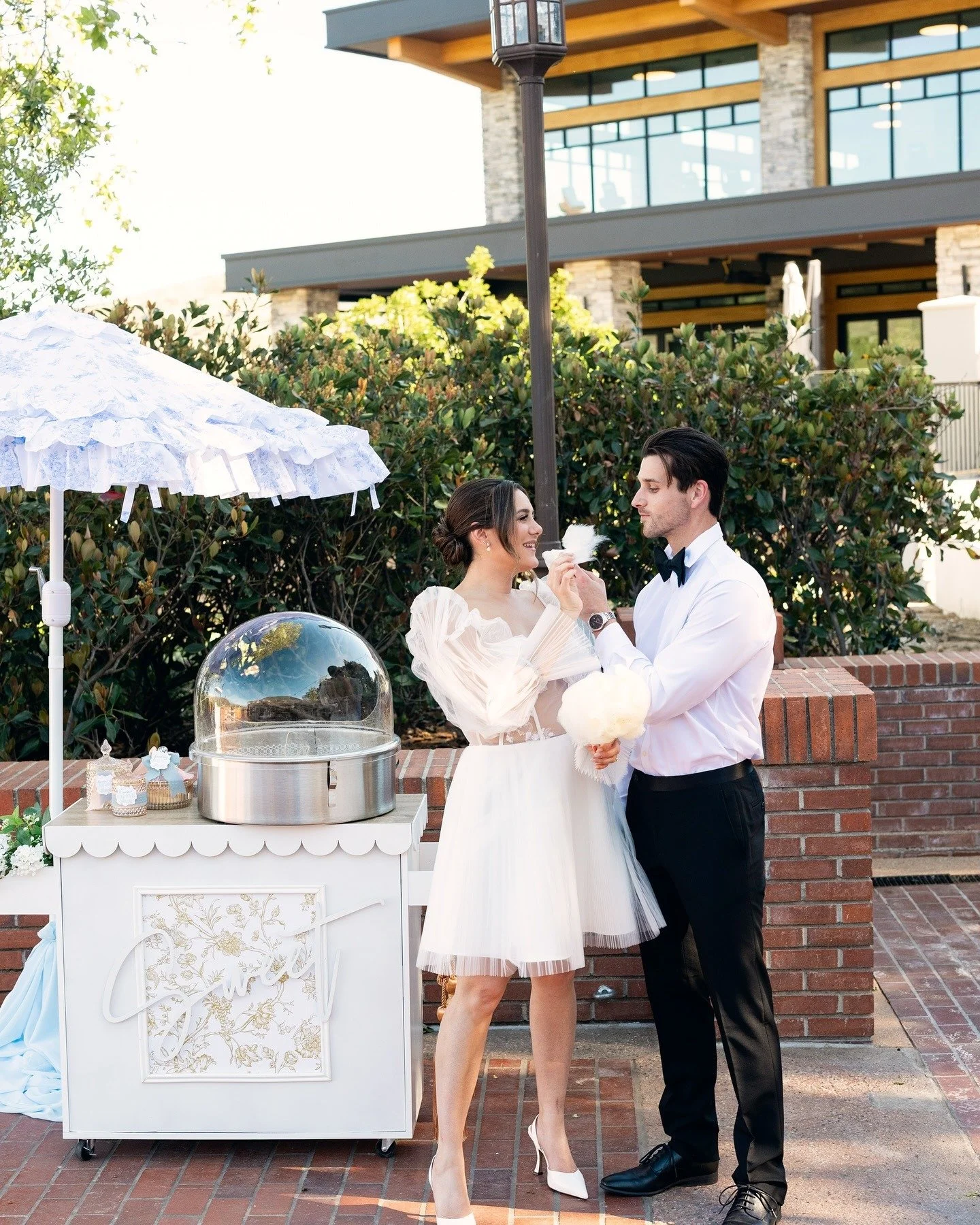 A bride and groom sharing cotton candy at their outdoor wedding reception, with a white cotton candy cart and decorative umbrella nearby.