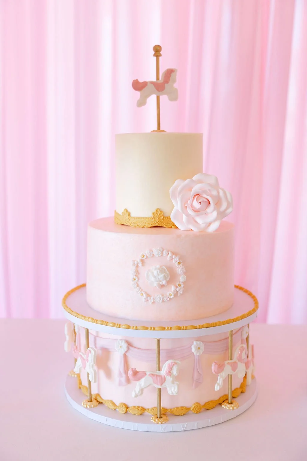 A pink carousel themed cake with two tiers, decorated with white and pink carousel horses, a large white flower, and a smaller pink flower. The cake is topped with a white cylindrical tier with a pink carousel horse topper. The background is pink cur