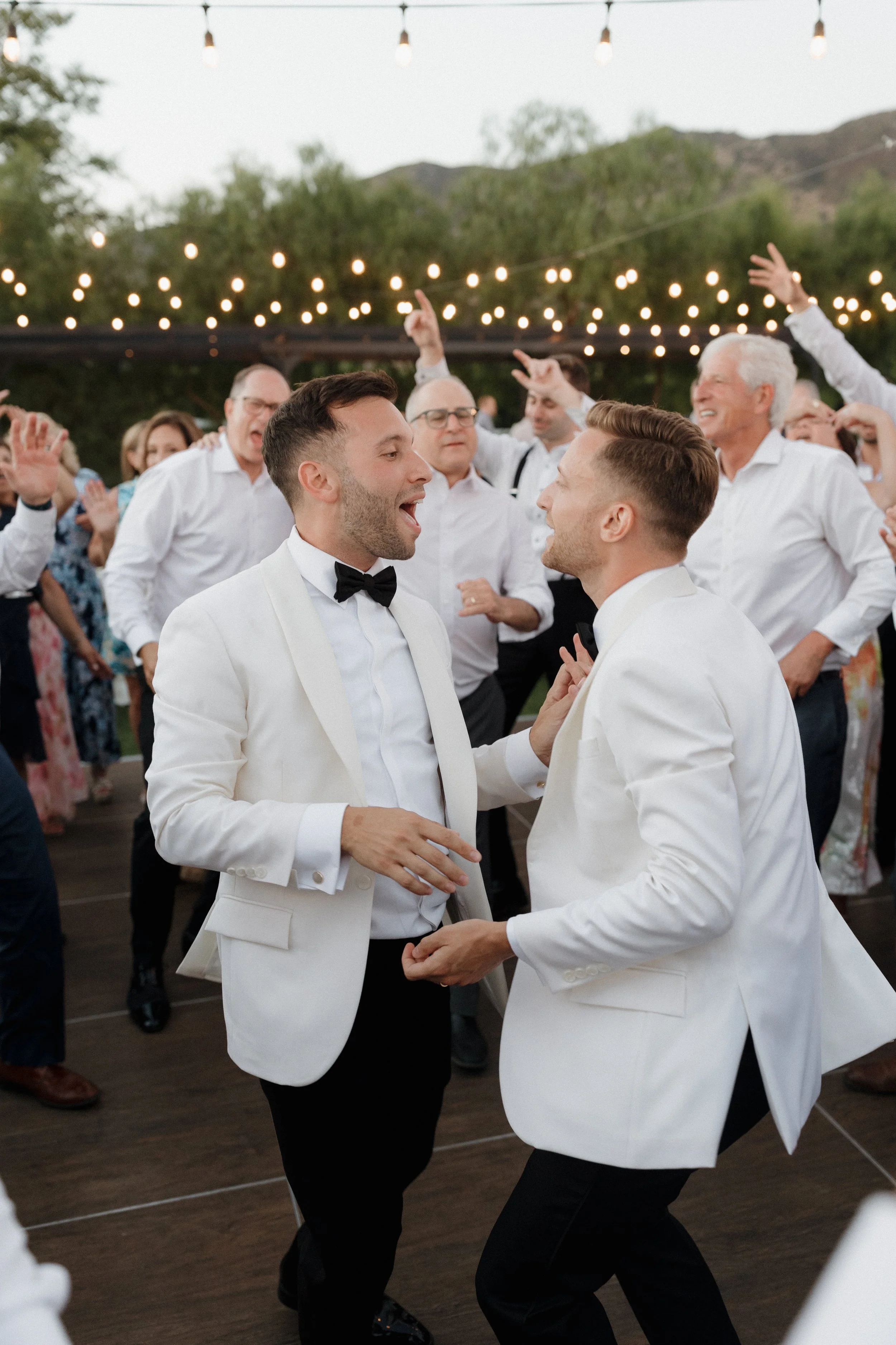 Two men in white tuxedos dancing and celebrating at a wedding reception outdoors, with guests and string lights in the background.