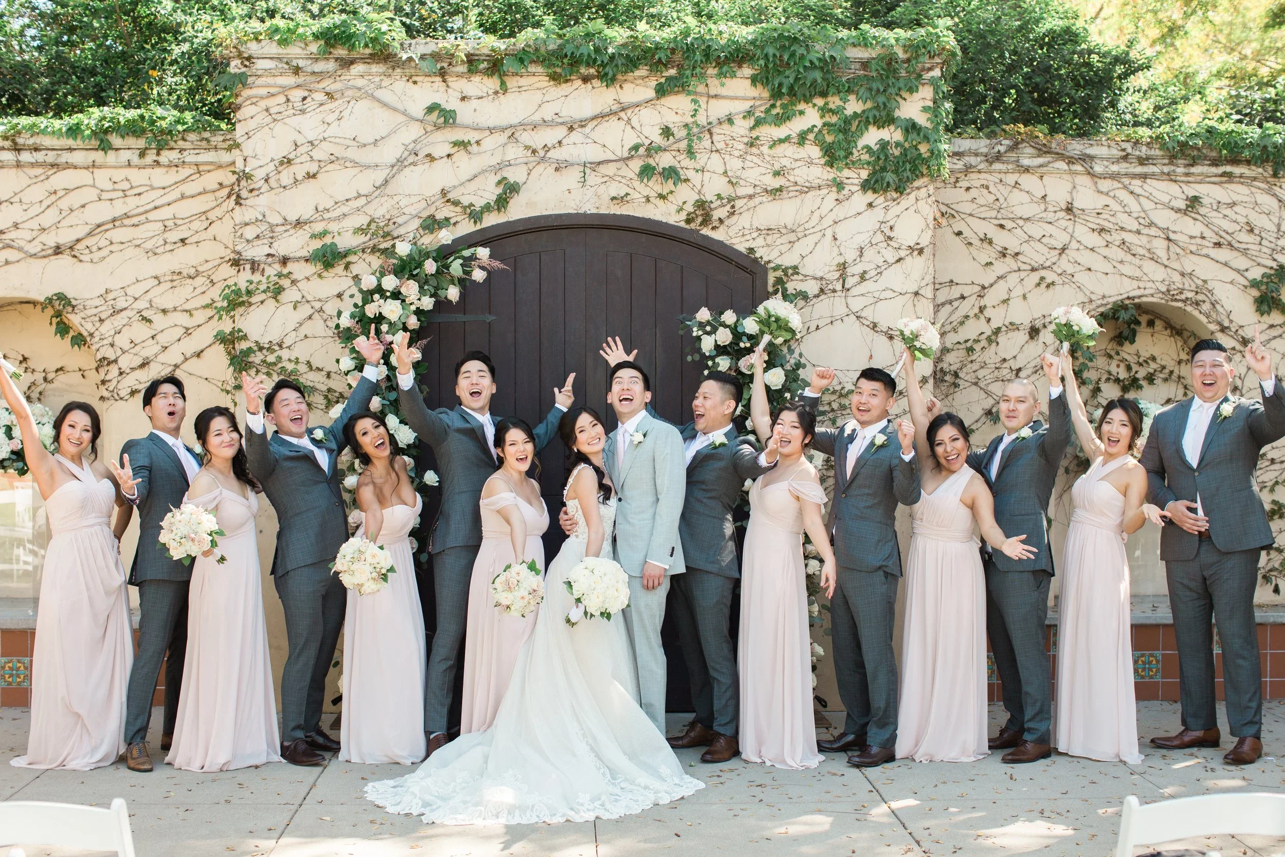 Group of wedding guests celebrating with a bride and groom in front of a rustic wall decorated with climbing vines and white flowers