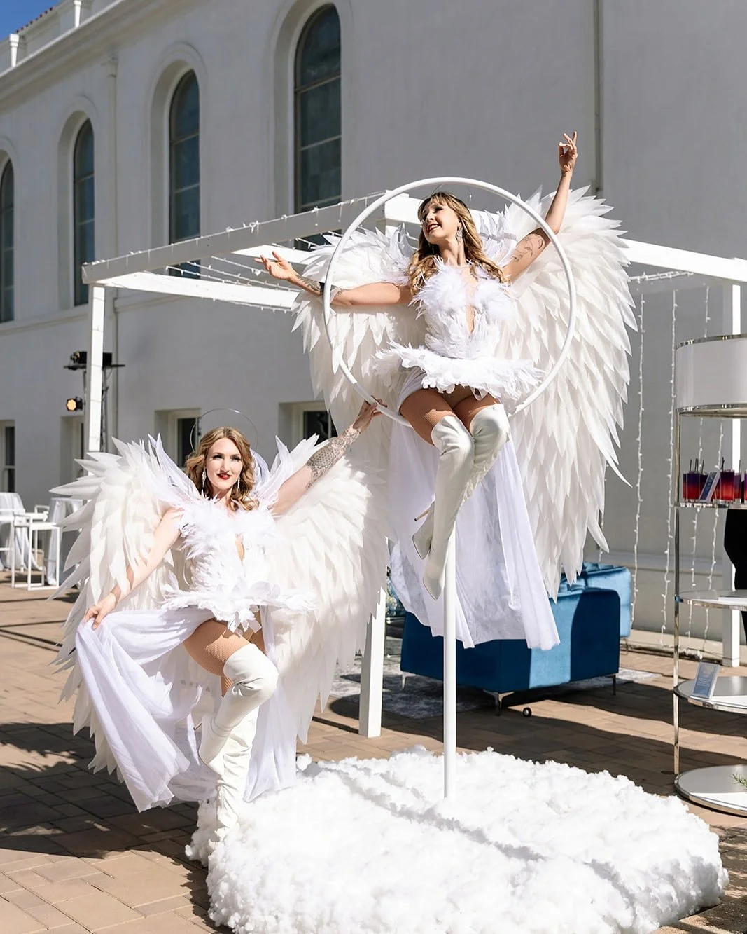 Two women dressed as angels with large white feathered wings and white costumes, one standing and one sitting on a white fluffy platform outdoors at a white building.