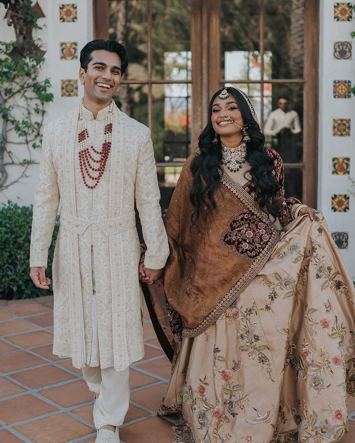 A man and woman dressed in traditional Indian wedding attire, holding hands and smiling, standing in front of a decorative wooden and tile background.